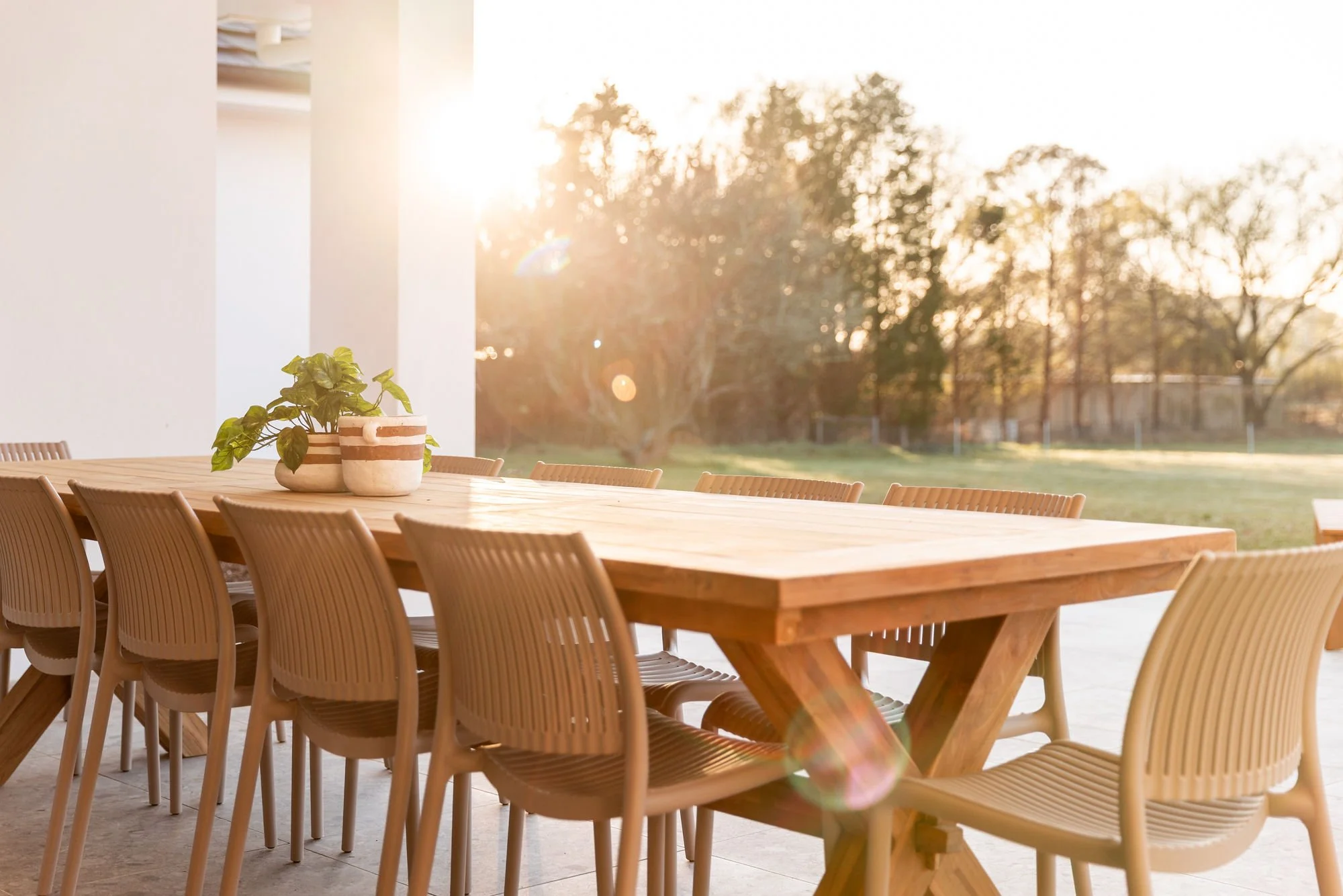 Empty outdoor dining table with beige chairs and potted plants, with sunlight in the background and trees outside.