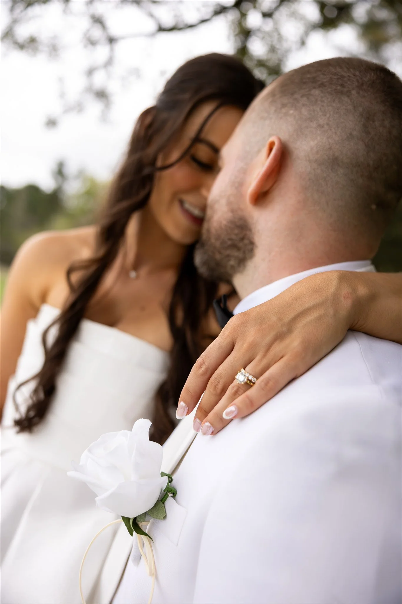 Close-up of a bride and groom embracing, the bride wearing a white dress and a wedding ring, and the groom in a white tuxedo with a white boutonniere, outdoors with trees in the background.