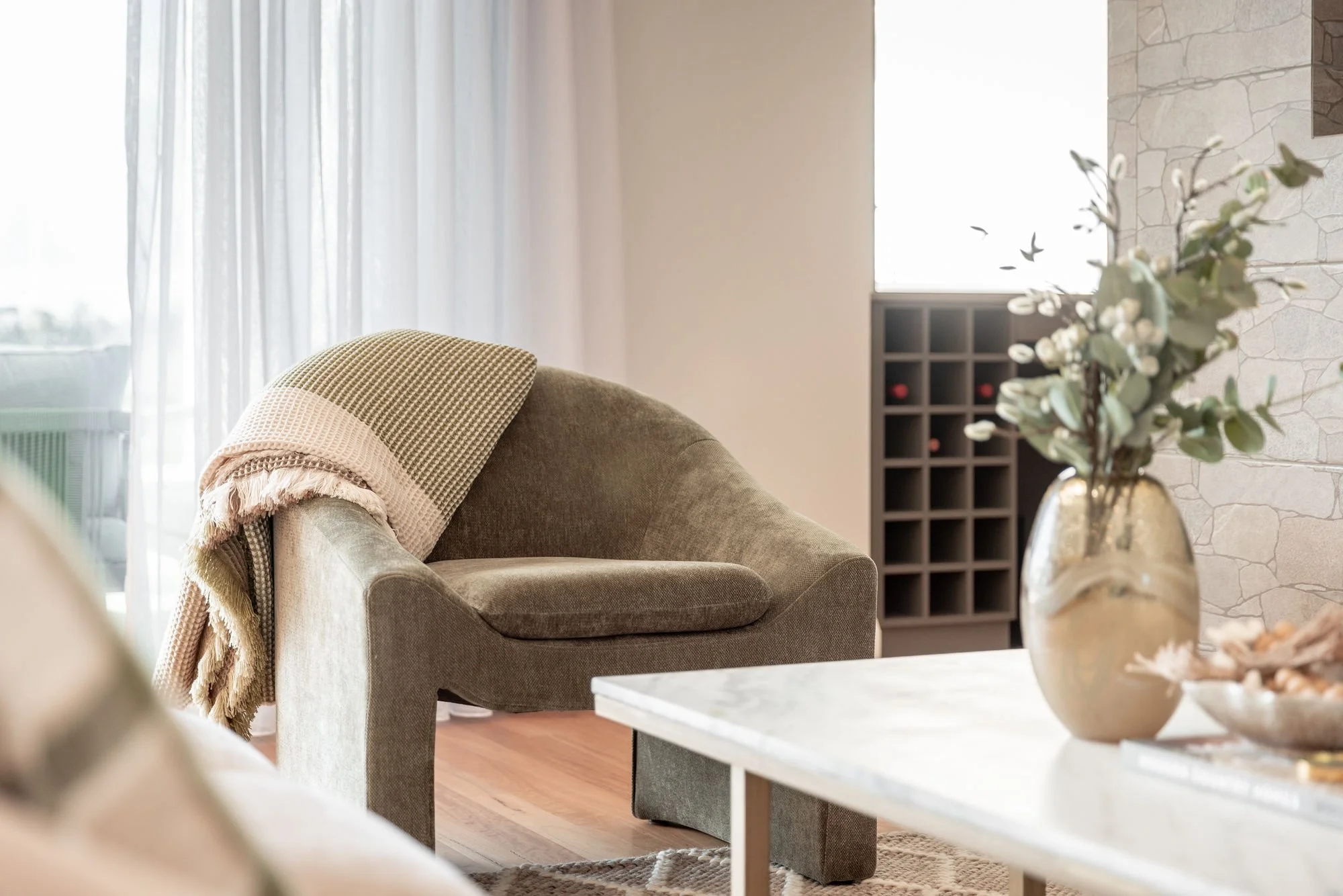 Living room with beige armchair and blanket, white curtains, stone accent wall, and vase with flowers on coffee table