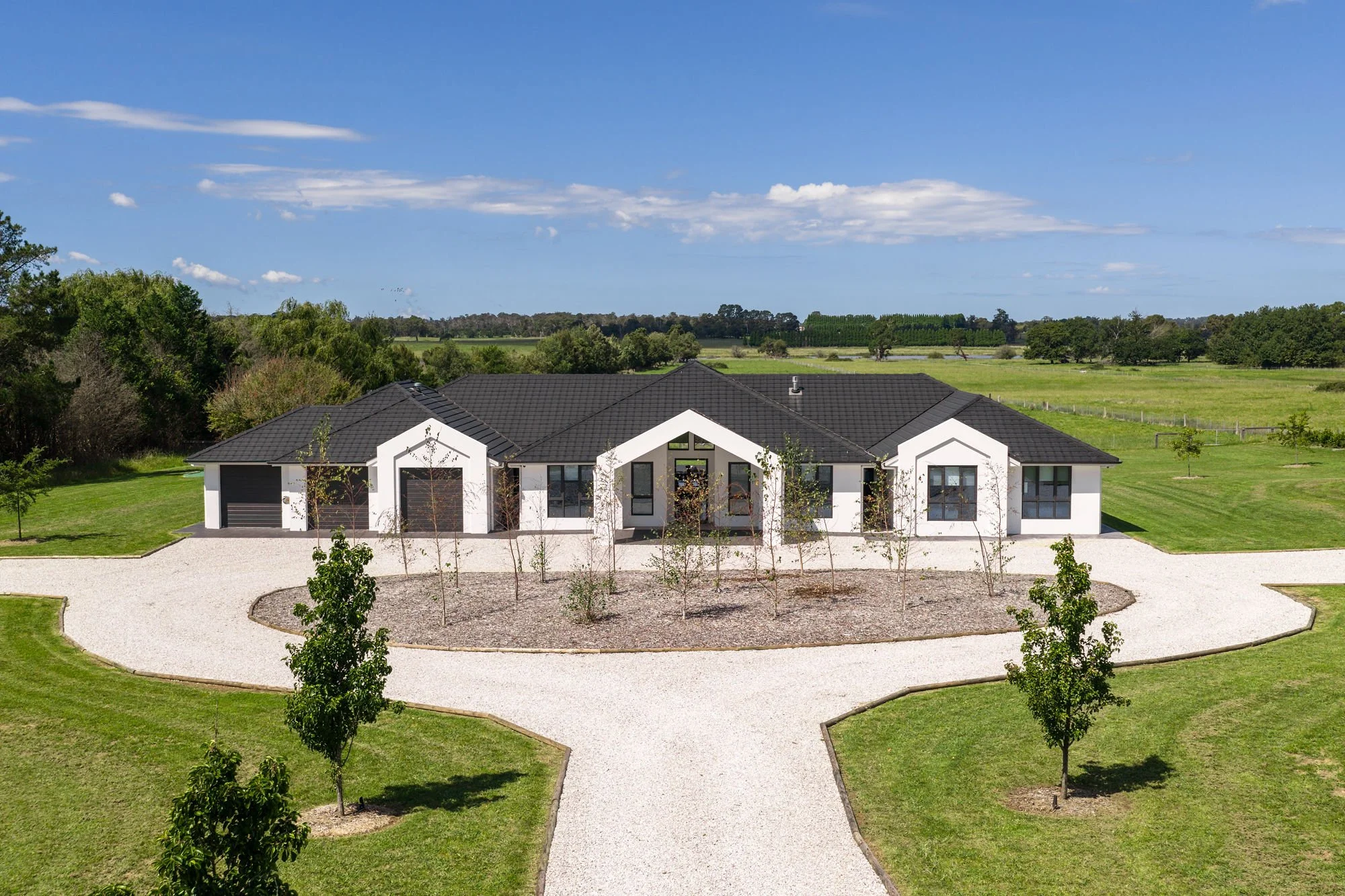 Modern single-story house with white exterior walls and a black roof, surrounded by a well-maintained lawn and a gravel driveway, across open fields and trees under a blue sky.