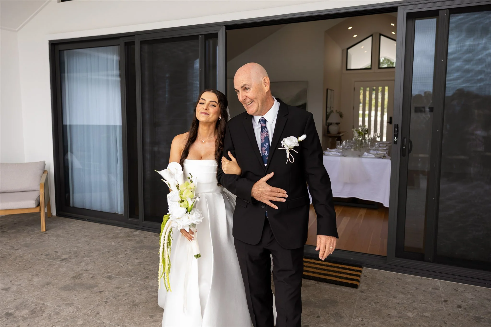 A bride walking arm-in-arm with her father through a doorway at a wedding reception. The bride is wearing a white wedding gown and holding a bouquet of white flowers. The father is dressed in a black suit with a boutonniere.