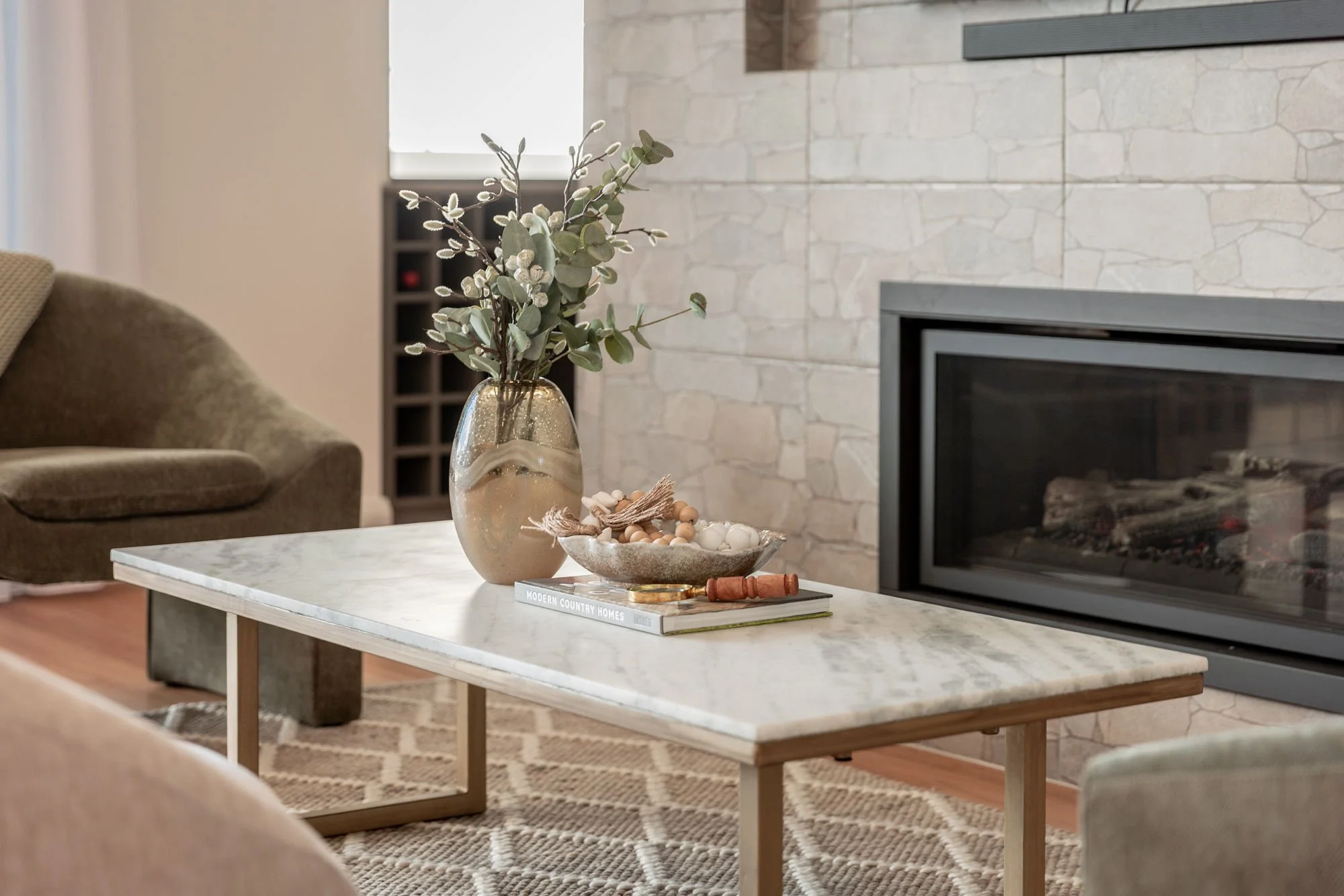Living room with a marble-top coffee table, decorative vase with green foliage, tray with decorative balls, book titled 'Modern Country Homes', and a fireplace with stone surround.
