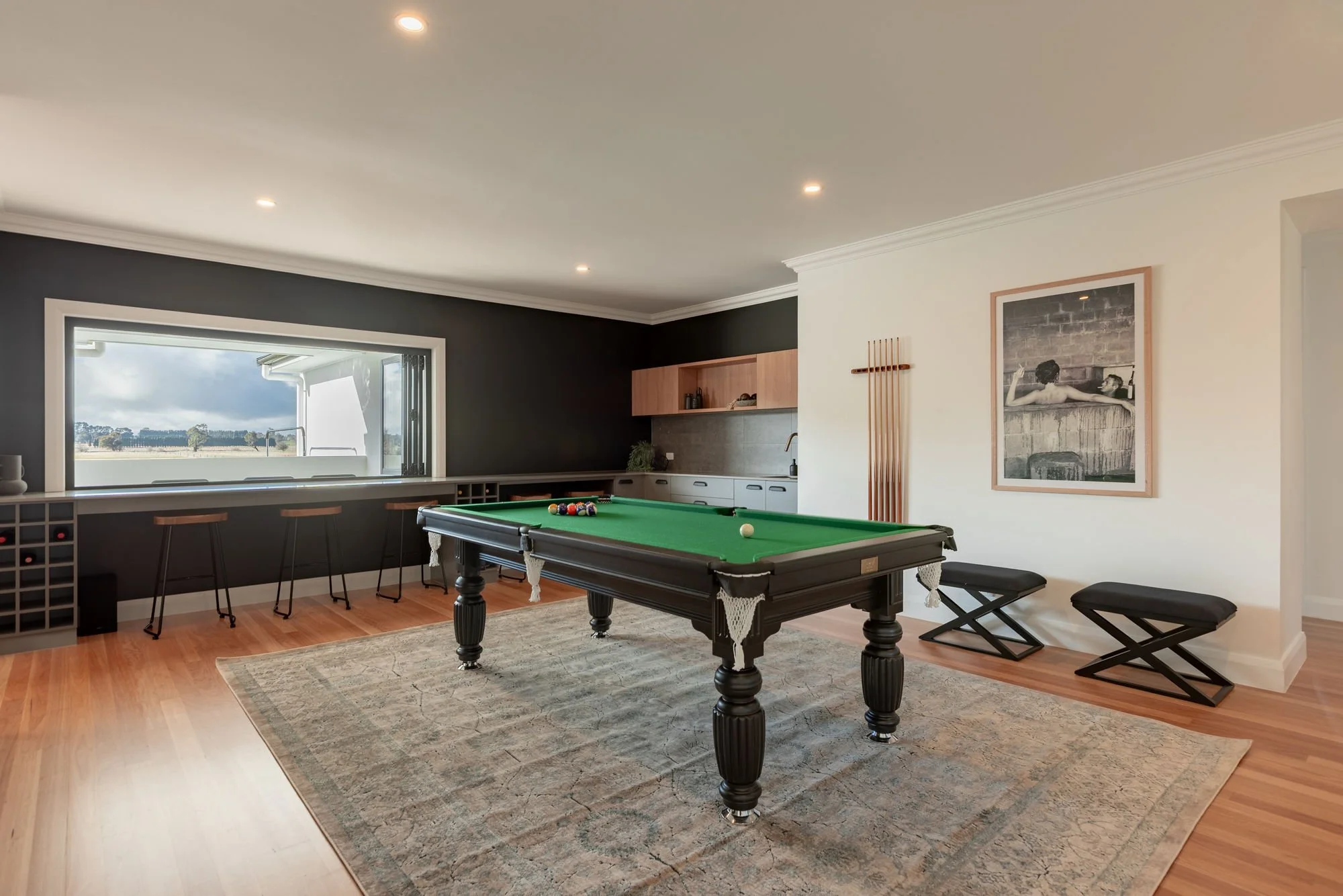 A game room with a black pool table, green felt, pool balls, and cue stick, situated on a patterned rug with benches and a window showing an outdoor view.
