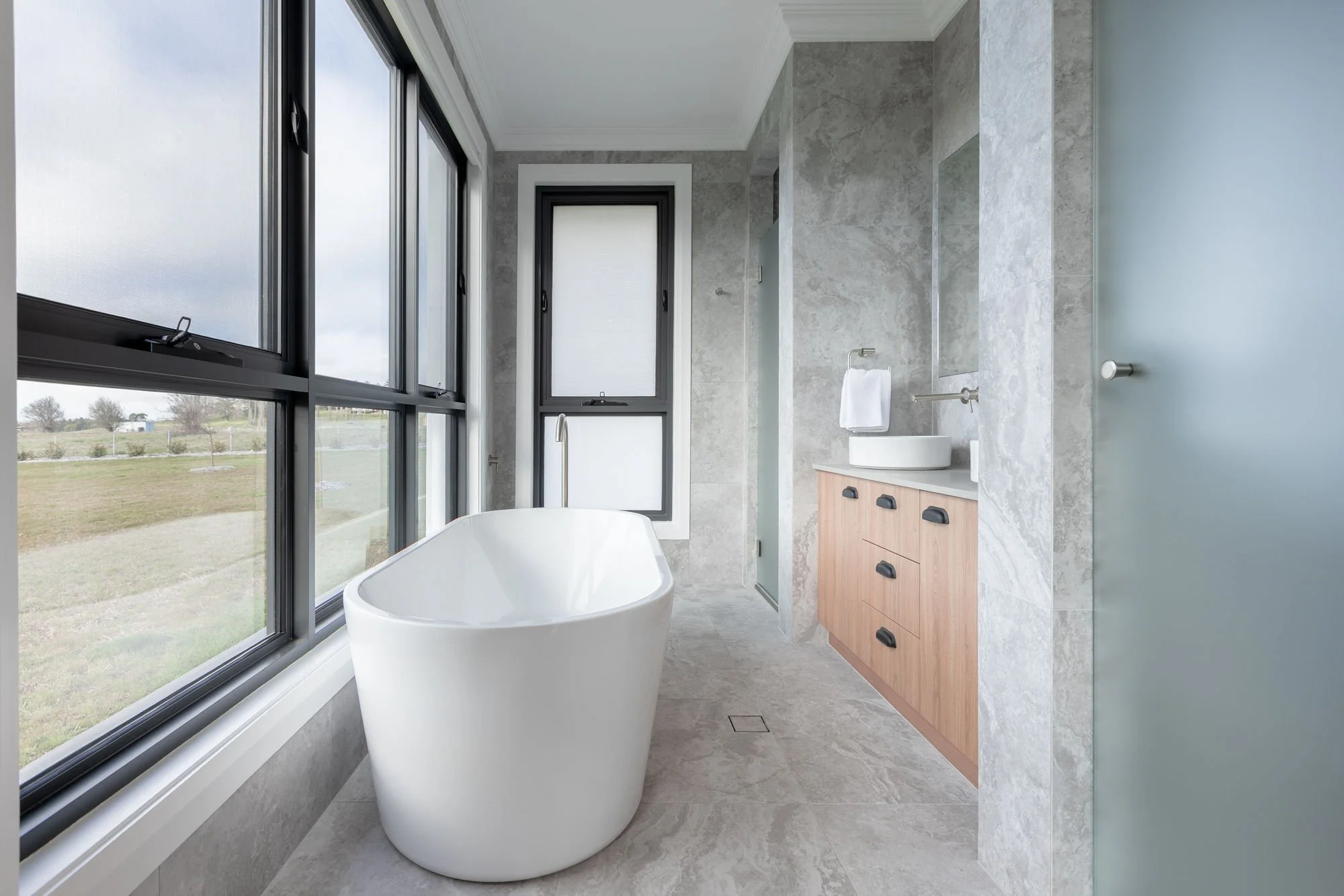 Modern bathroom with large windows, free-standing soaking tub, wooden vanity with black handles, round vessel sink, and gray marble tiles.