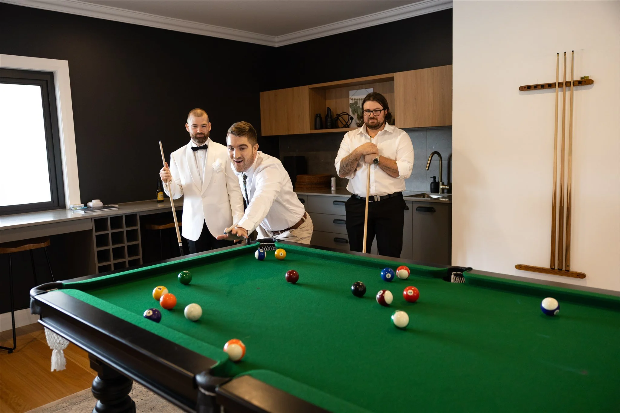 Three men dressed in formal attire playing billiards in a modern room with dark and light walls, pool balls scattered on the green felt table, and pool cues hanging on the wall.