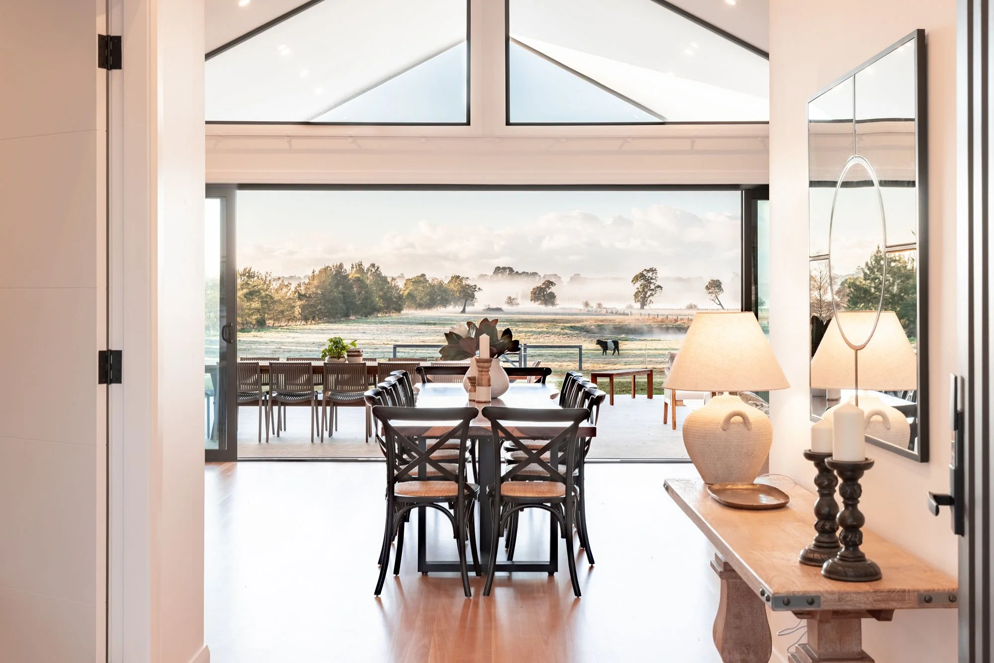 View of a dining area with a large glass sliding door leading to an outdoor patio, overlooking a scenic field with trees and a horse in the distance. Inside, there is a wooden table with black chairs, a console table with lamps, and a mirror, bathed 