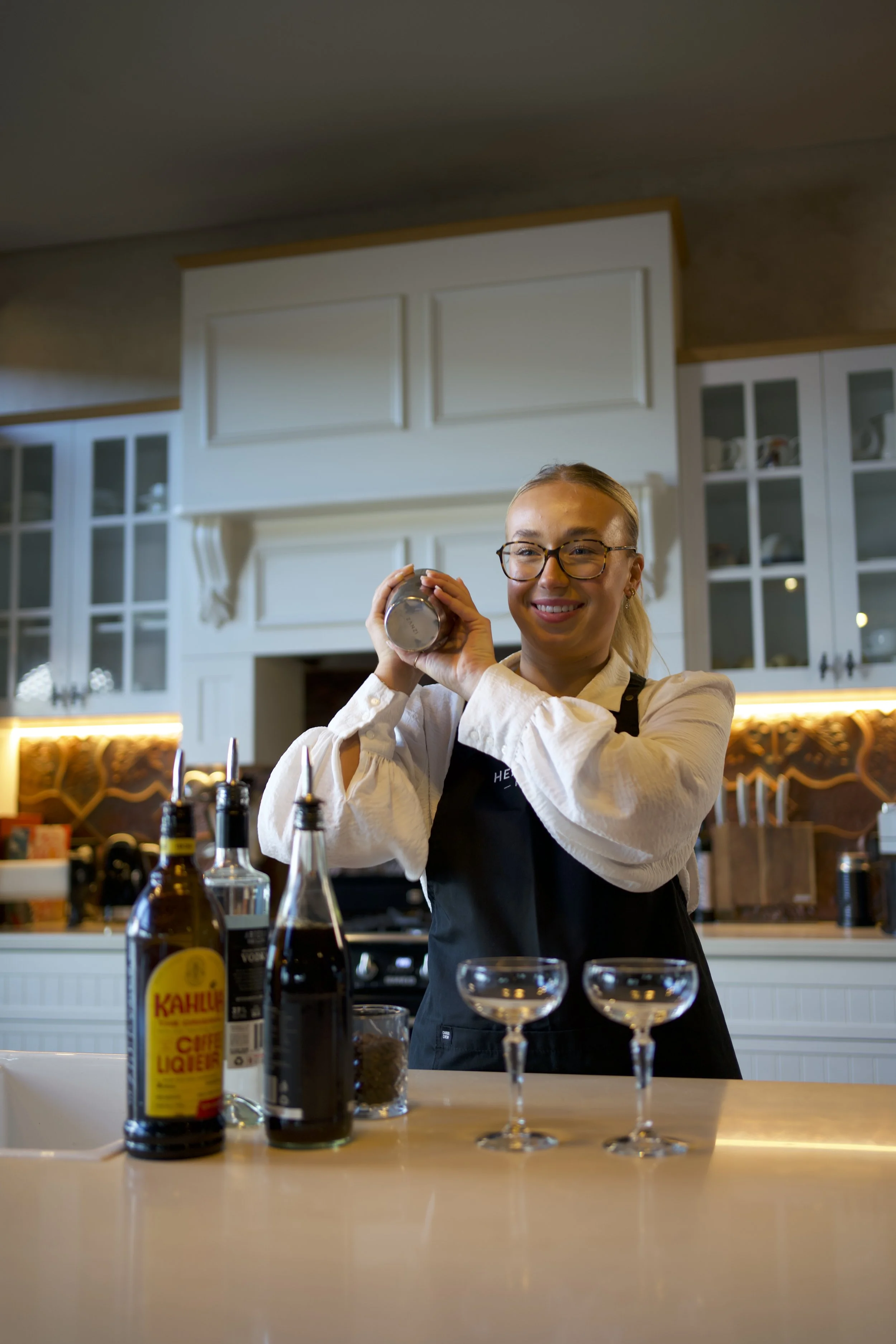 A woman smiling in a kitchen, holding a cocktail shaker, with liquor bottles and glasses on the counter.