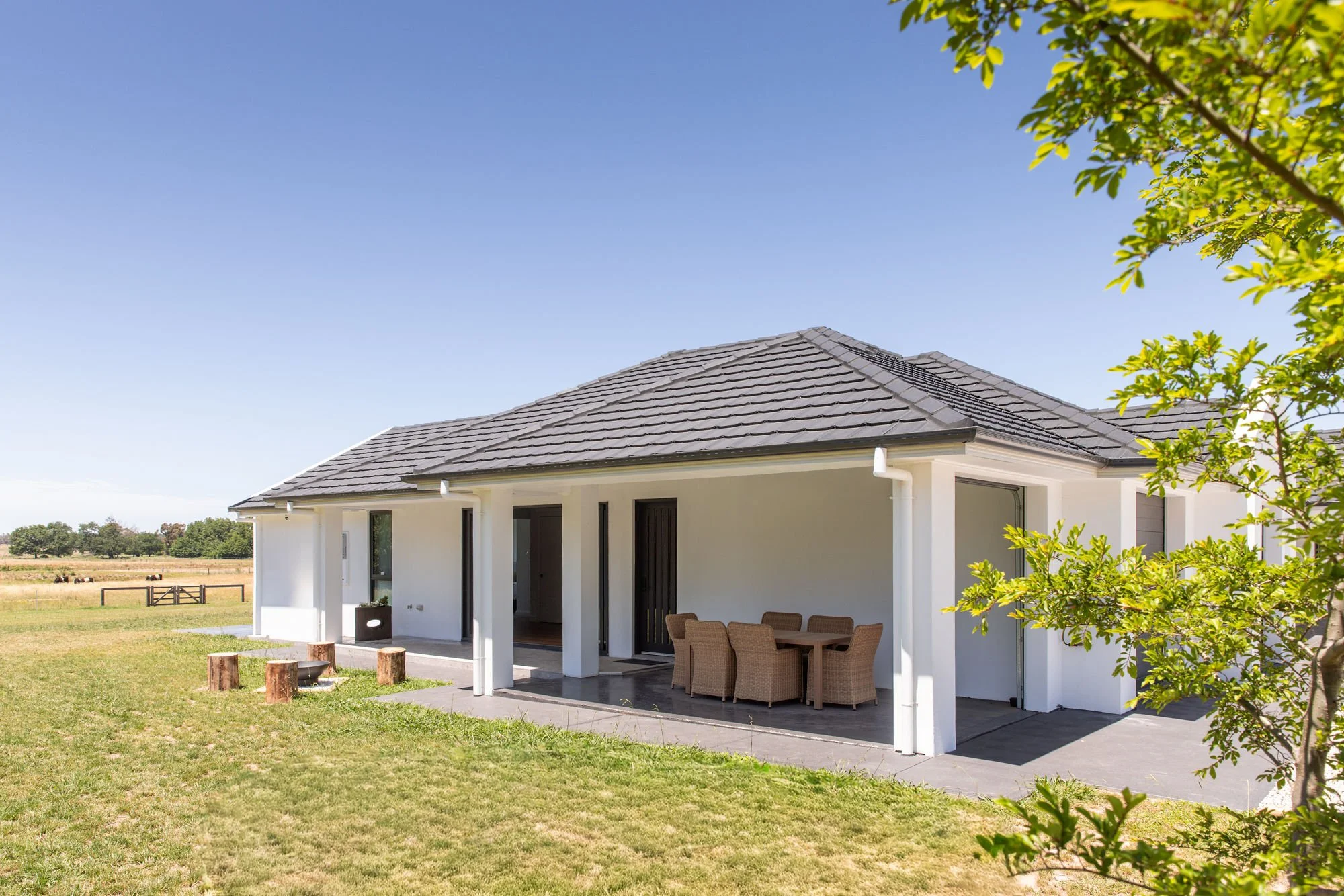 A modern single-story house with a gray tiled roof, white exterior walls, and an outdoor patio with four wicker chairs and a table, set in a grassy yard under a clear blue sky with a few trees and a distant fence.