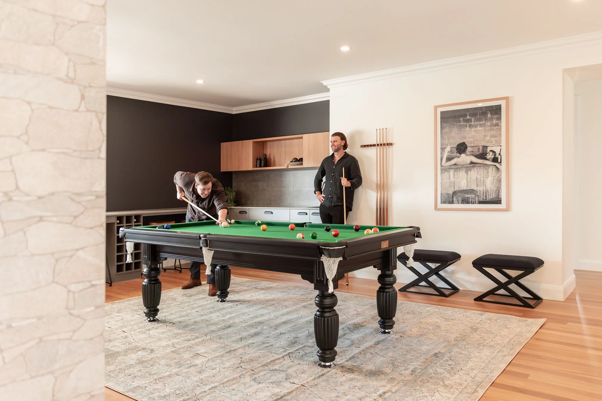 Two men playing pool in a modern living room with a black and white photo on the wall, a wooden shelf, and a beige rug.