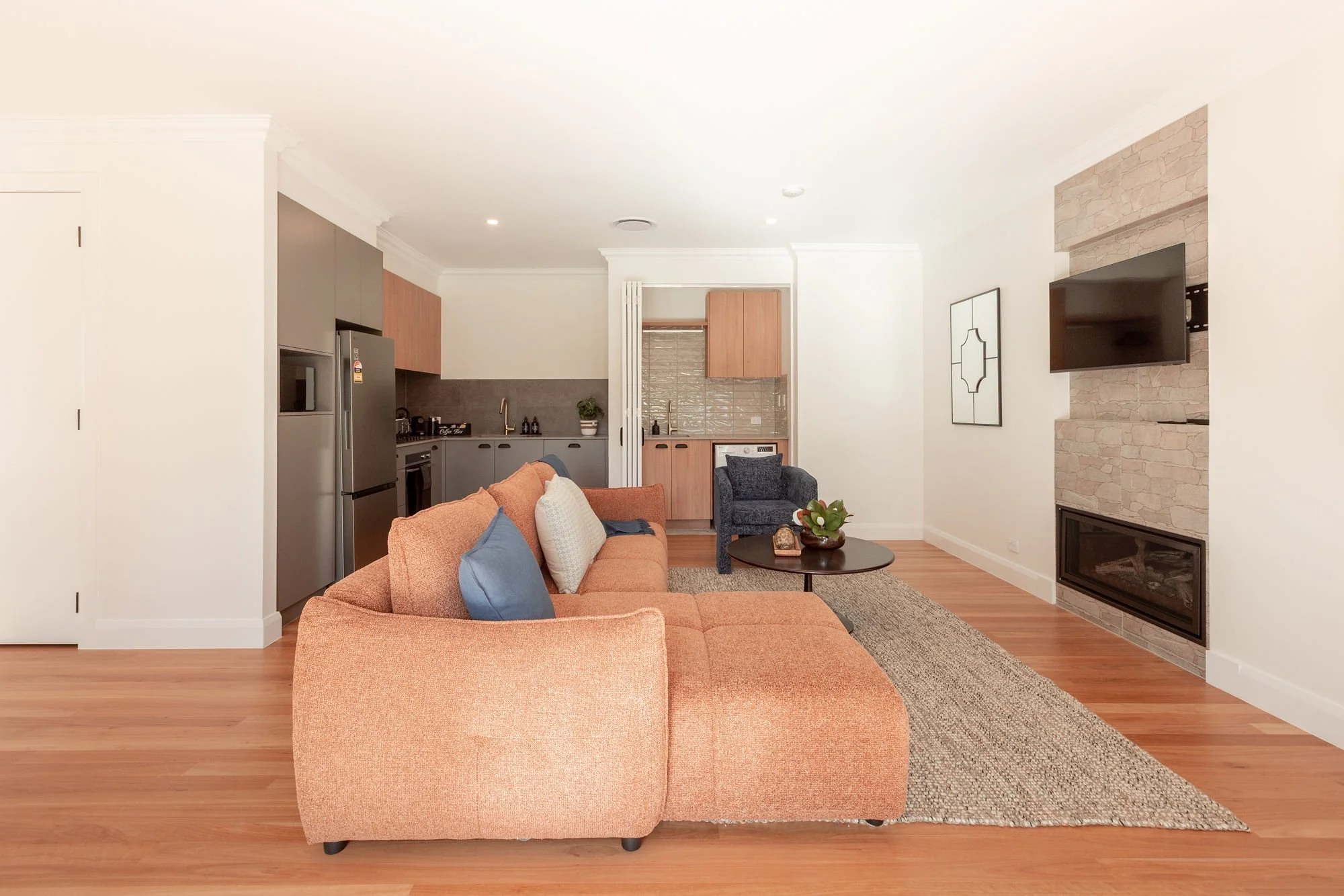 A spacious living room with a peach-colored sofa, blue and white pillows, a round coffee table with a plant, and a TV mounted on a stone fireplace. The background includes a kitchen area with gray and wooden cabinets.