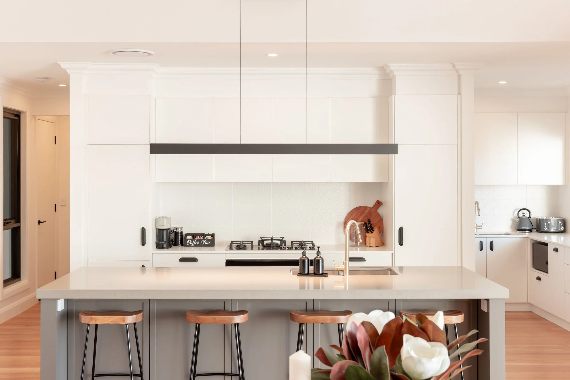 Modern white kitchen with a large island, wooden bar stools, and built-in appliances.