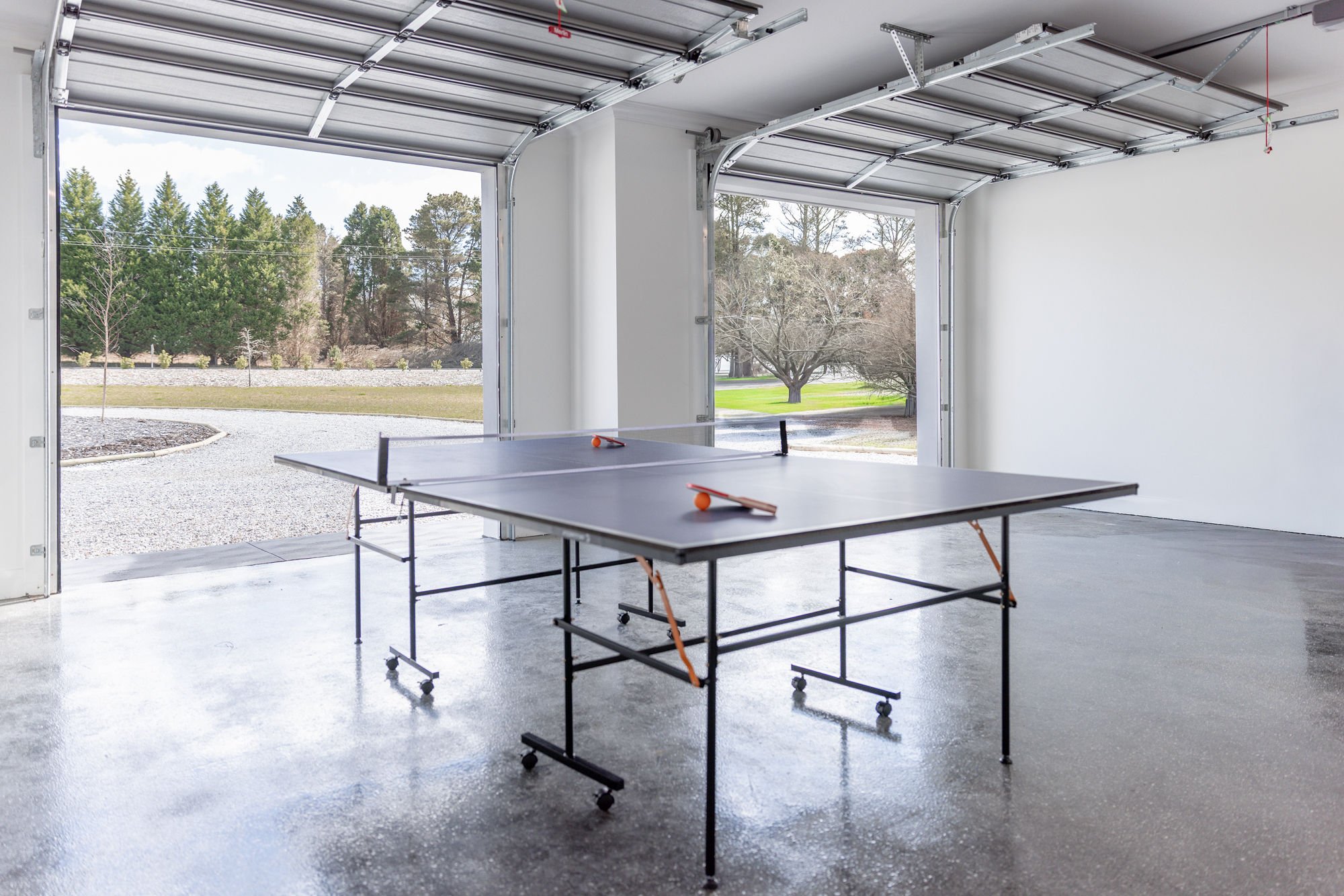 Garage with an open door showing a ping pong table set up inside, with paddles and balls on it, and trees visible outside.