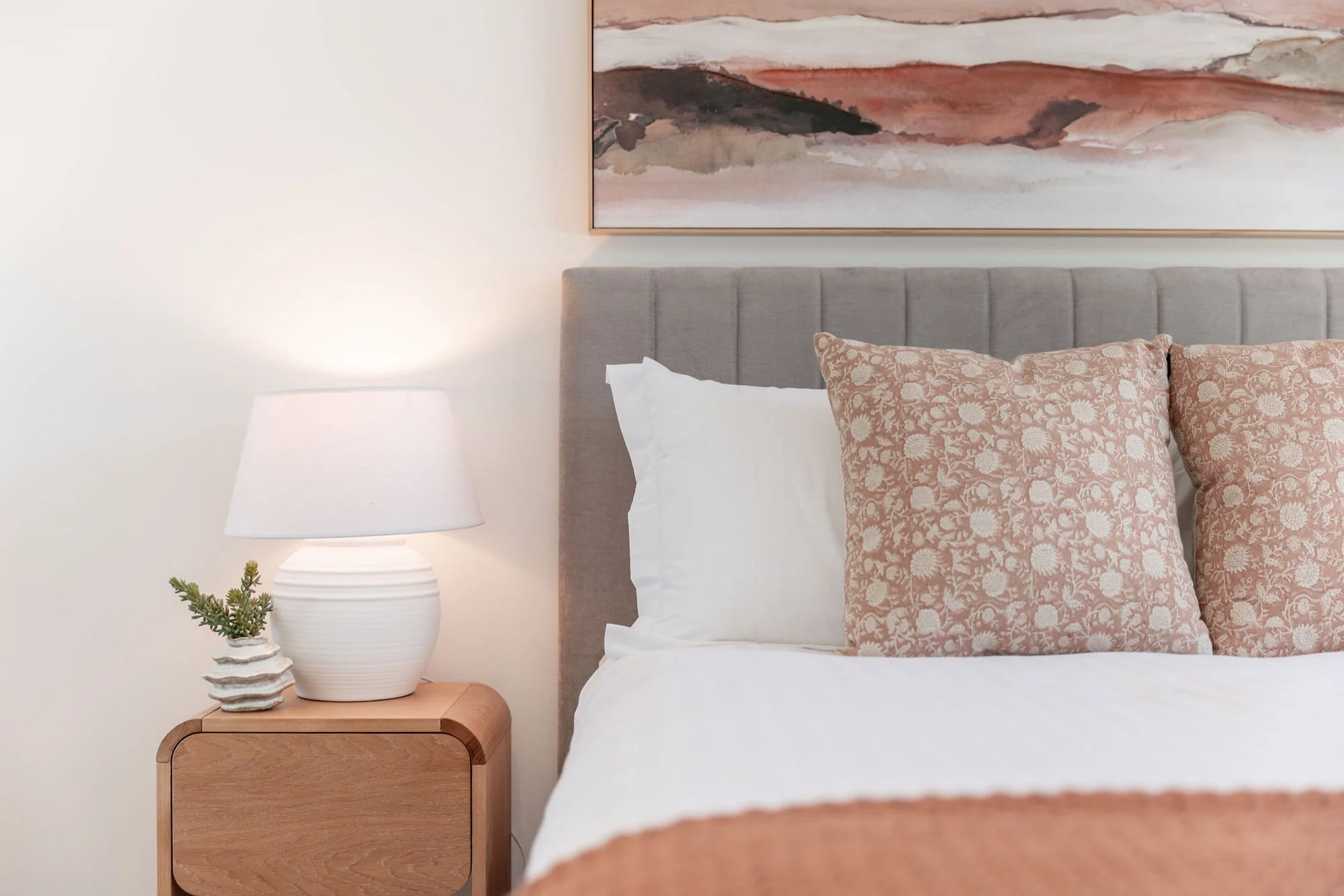 A neatly made bed with a beige upholstered headboard, two white pillows, and two patterned throw pillows in shades of pink and beige. To the left, a wooden nightstand holds a white ceramic table lamp and a small decorative pot with greenery. A framed