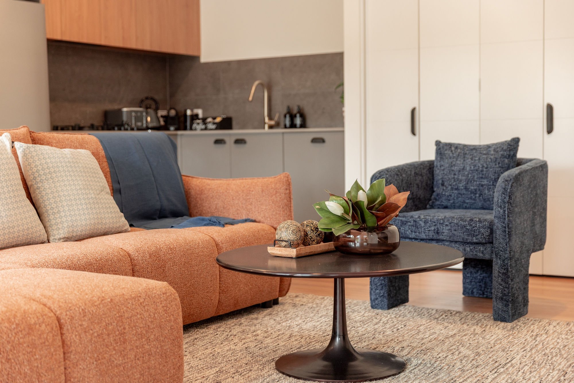 Living room with a peach sofa, a dark blue armchair, a coffee table with a plant and decorative objects, and a kitchen in the background.