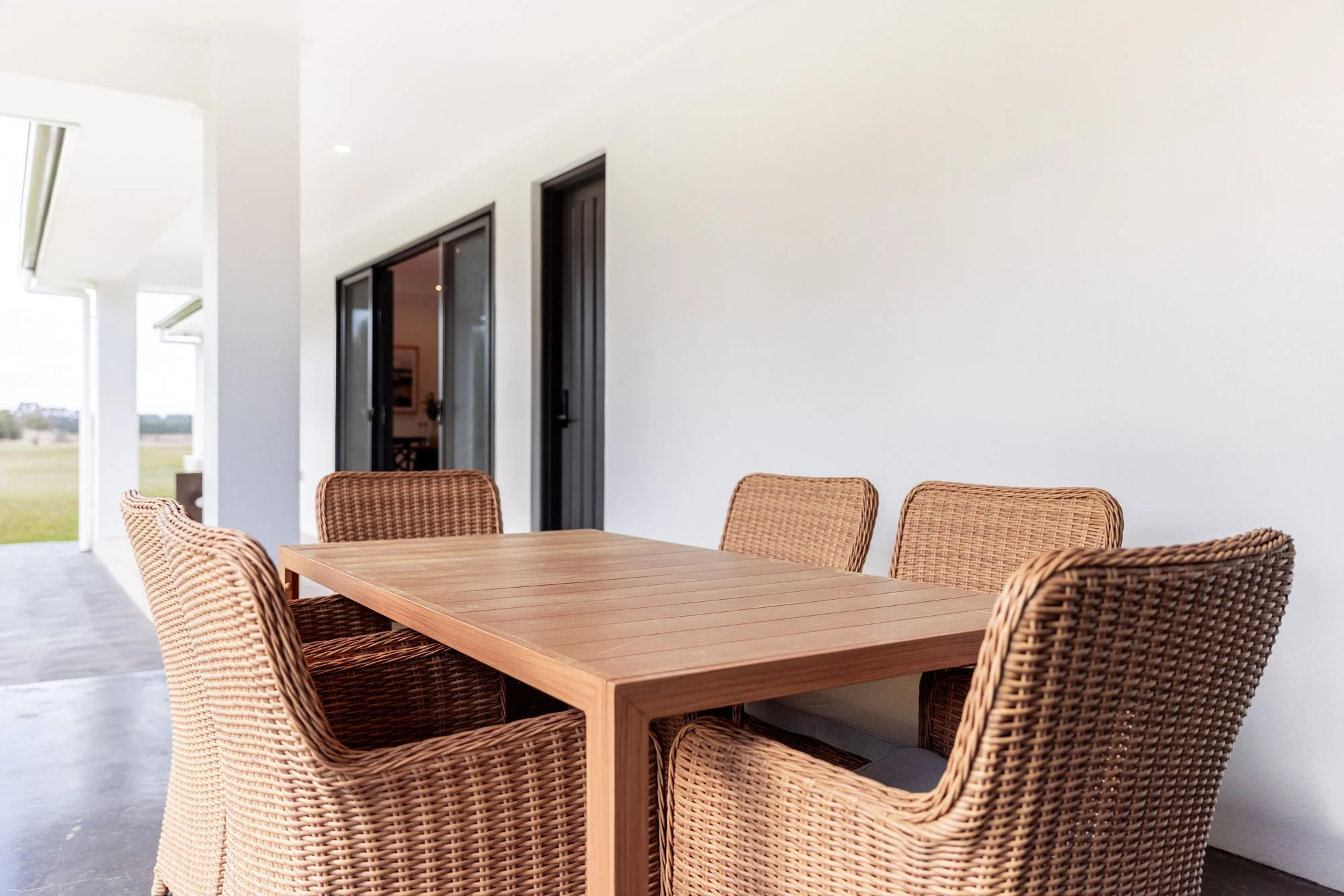 Outdoor patio with a wooden table and six wicker chairs, white building wall, sliding glass doors, and a view of a grassy field in the distance.