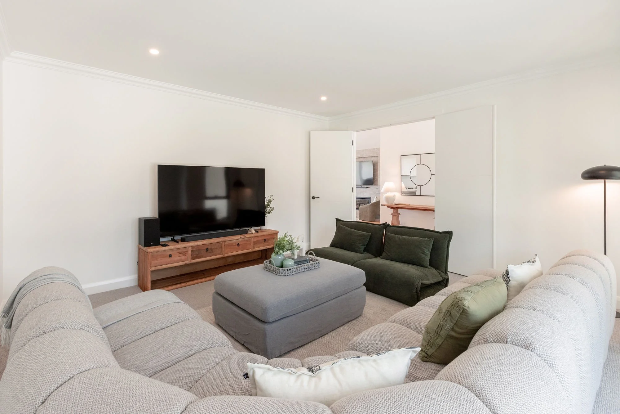 Living room with cream-colored sectional sofa, gray ottoman, dark green loveseat, flat-screen TV on wooden stand, and a standing lamp.