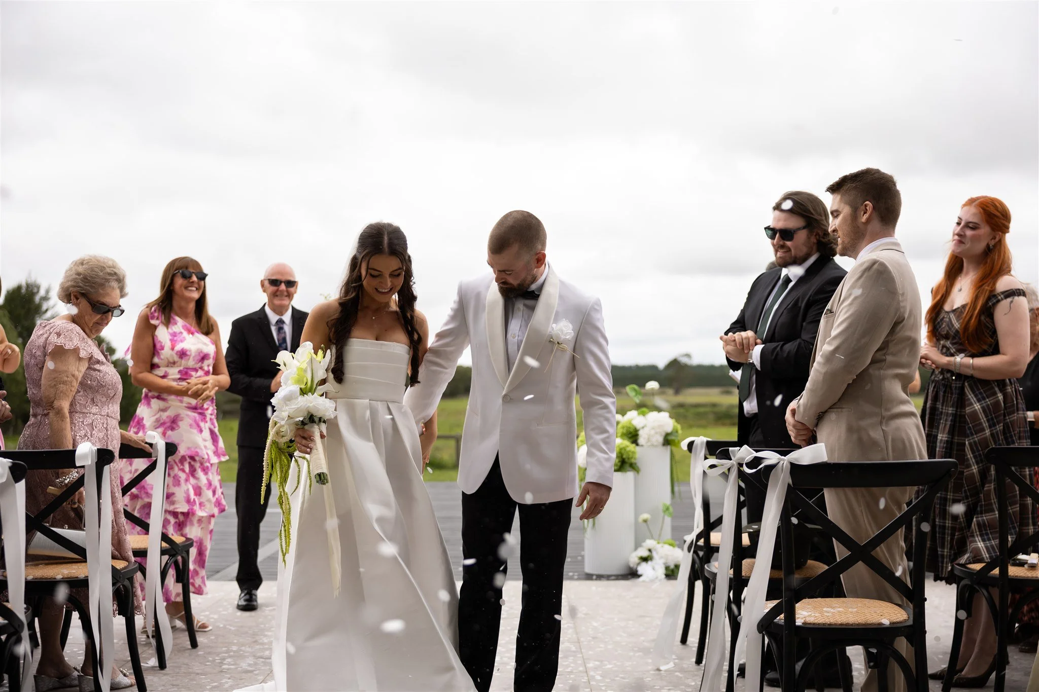 Bride and groom holding hands and walking down the aisle during an outdoor wedding ceremony, with guests standing on either side, some clapping and smiling.