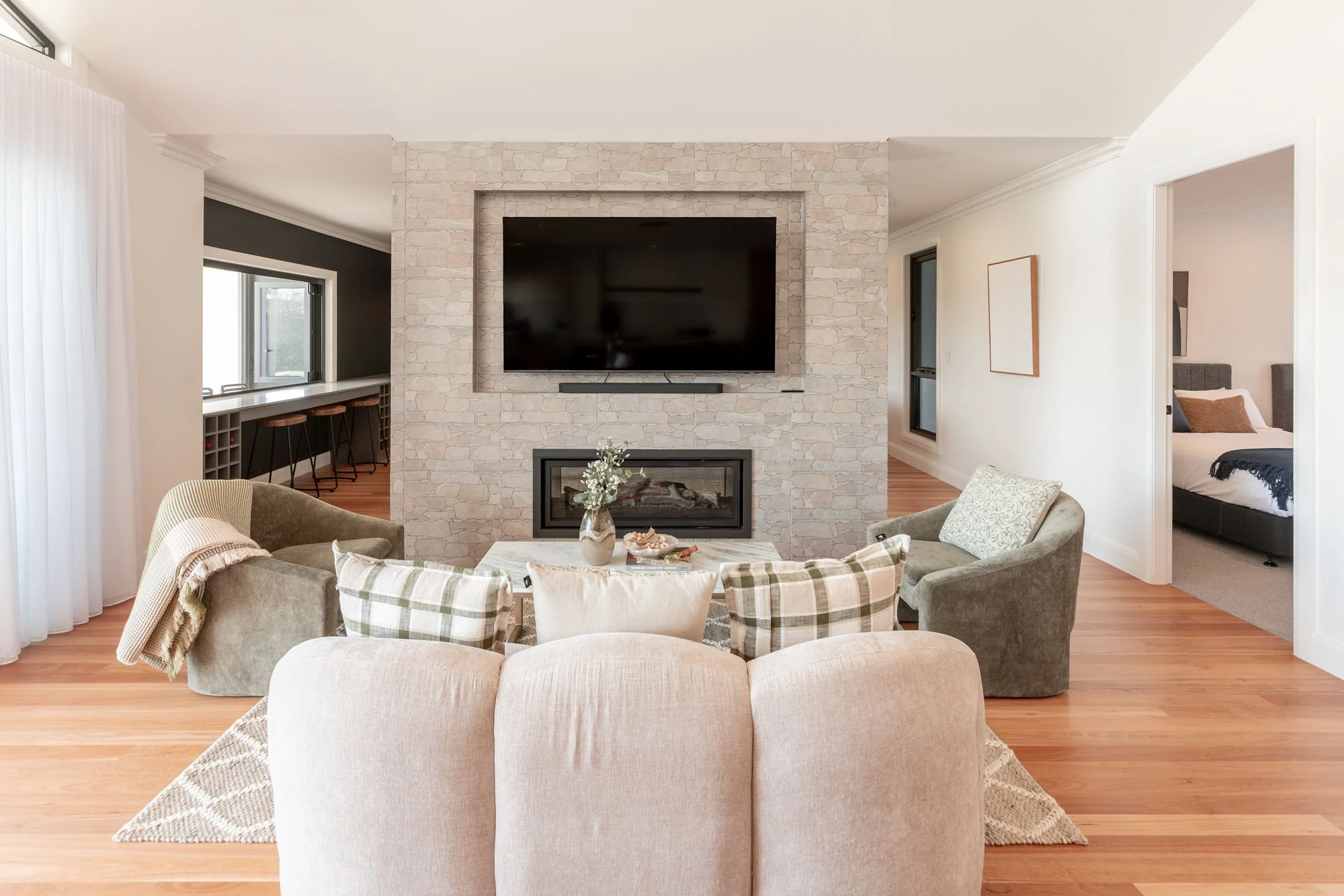 Living room with a fireplace, mounted TV, and seating including a sofa and armchairs, with hardwood floors and a view into a bedroom in the background.