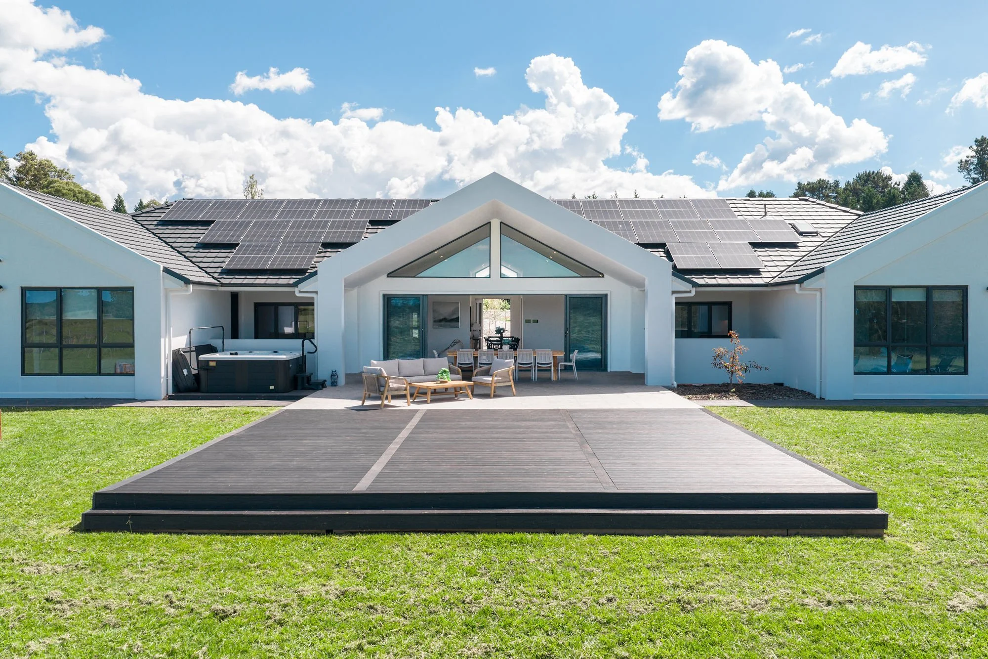 Modern white house with solar panels on the roof, large glass doors, a wooden deck with outdoor furniture, and a hot tub, set on a green lawn under a partly cloudy sky.