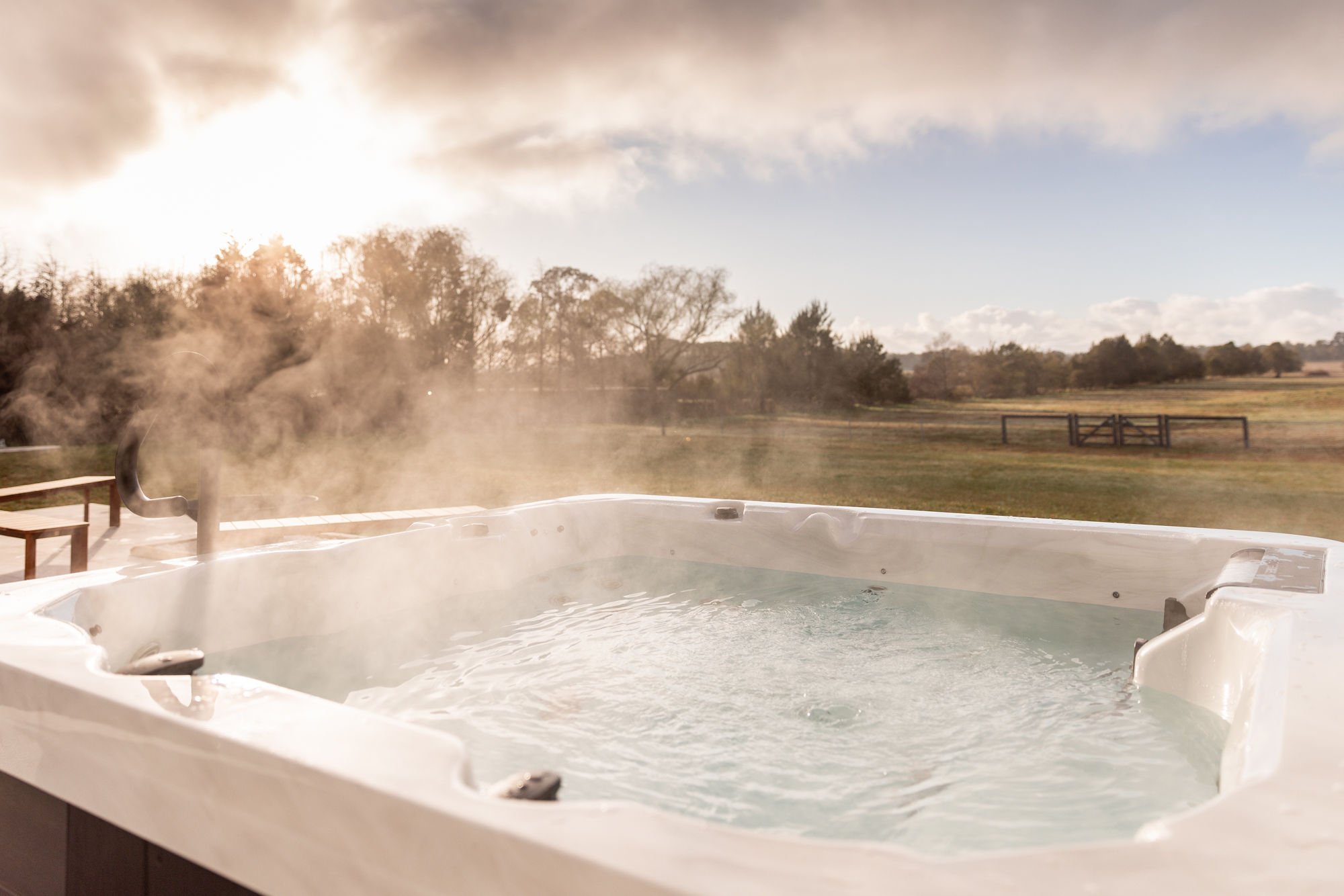 An outdoor hot tub filled with steaming water on a wooden deck, overlooking a grassy field with trees and a wooden fence in the background, during sunrise or sunset.