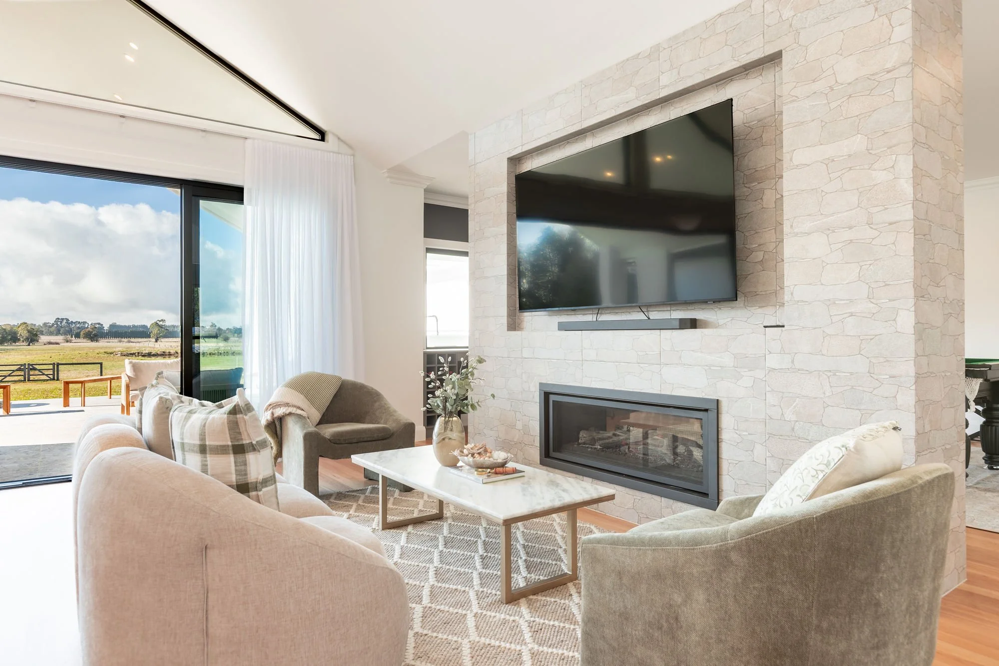 Living room with beige and gray furniture, a marble coffee table, a large wall-mounted flat-screen TV, a stone fireplace below the TV, and sliding glass doors opening to a outdoor view of fields and trees.