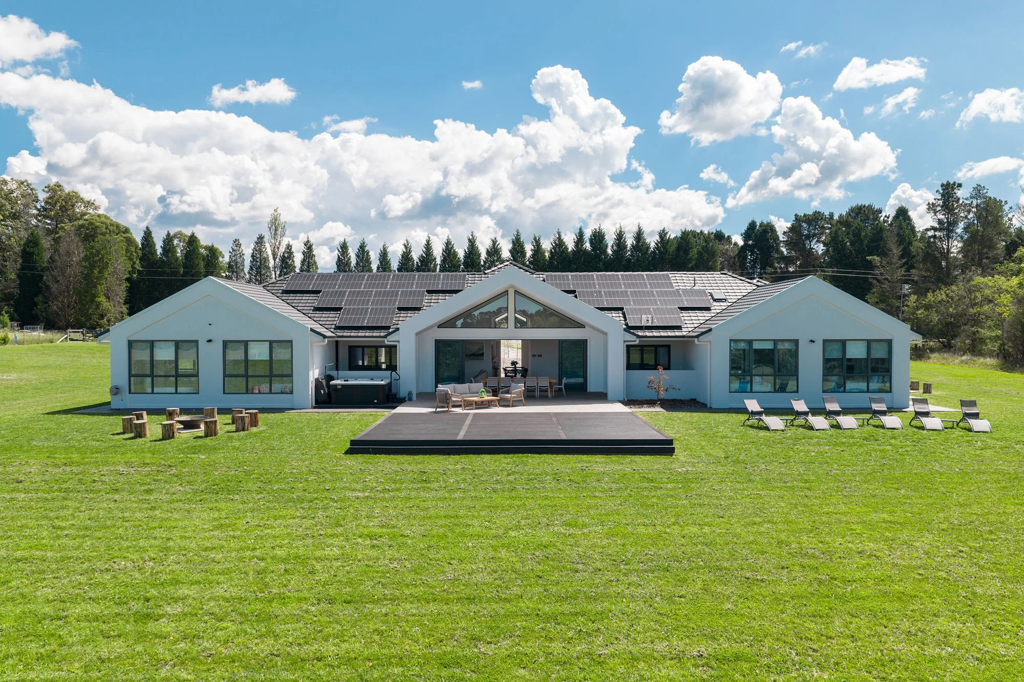 Modern house with solar panels on the roof, large windows, an outdoor patio, and lawn chairs on a grassy yard under a partly cloudy sky.