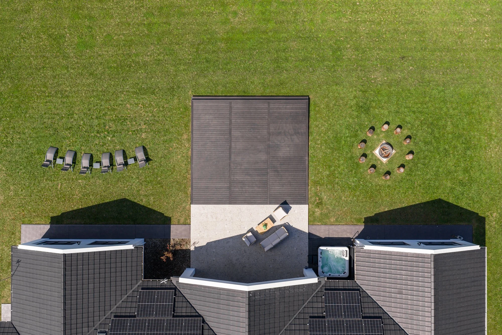 Aerial view of a house with solar panels on the roof, a patio with outdoor furniture, a hot tub, a grassy yard with chairs, and a fire pit with people around it.