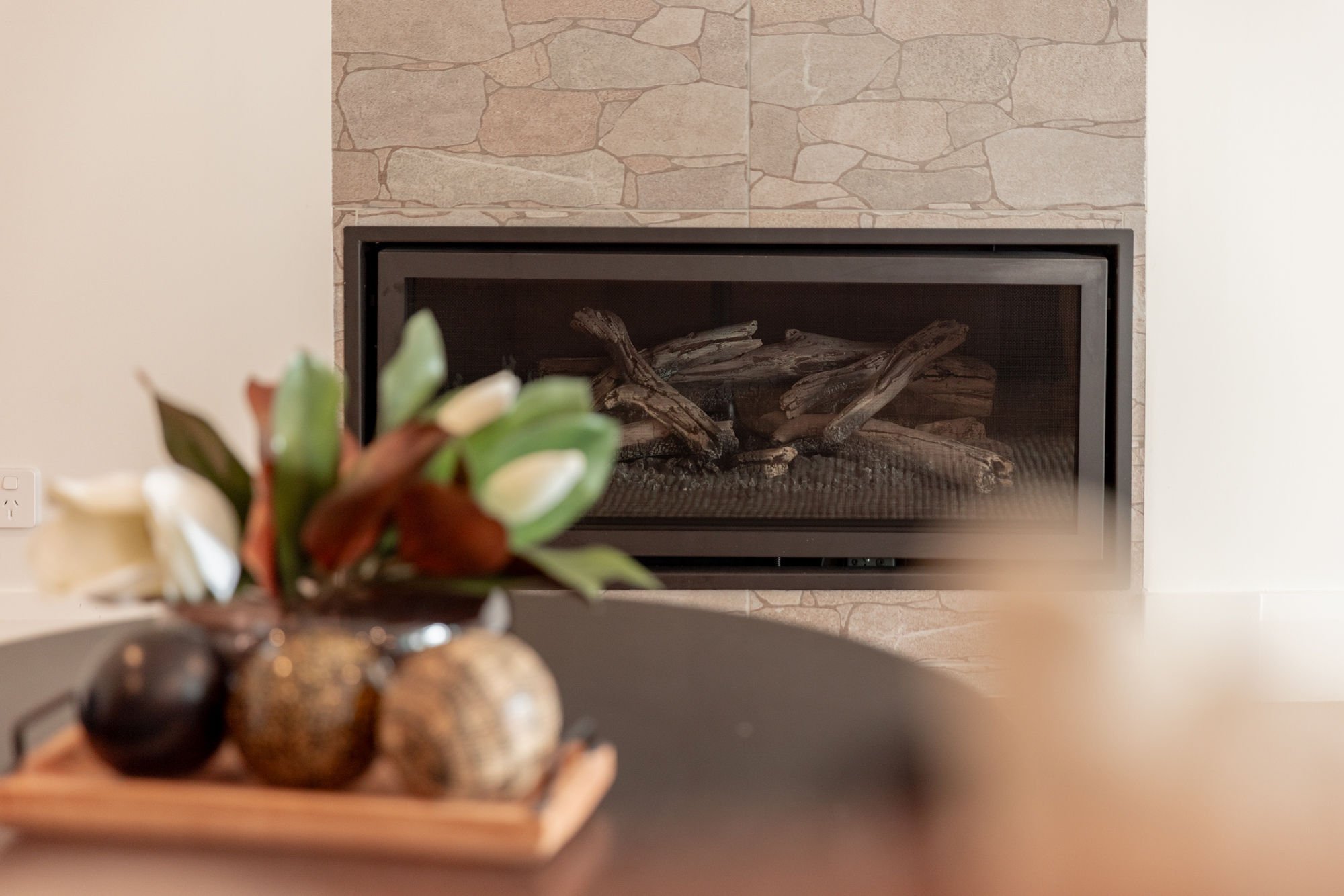 A fireplace with a stone surround and a black metal frame, containing logs inside. A blurred flower arrangement with white flowers and green leaves is in the foreground, out of focus.