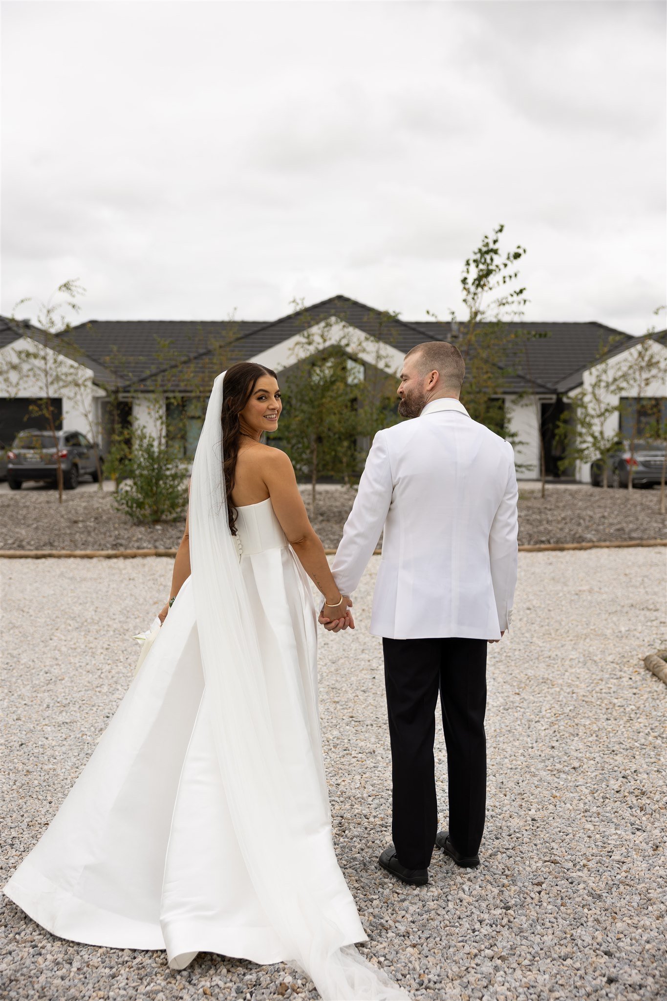 A bride and groom holding hands, dressed in wedding attire, standing outdoors on a gravel area, with houses and trees in the background.