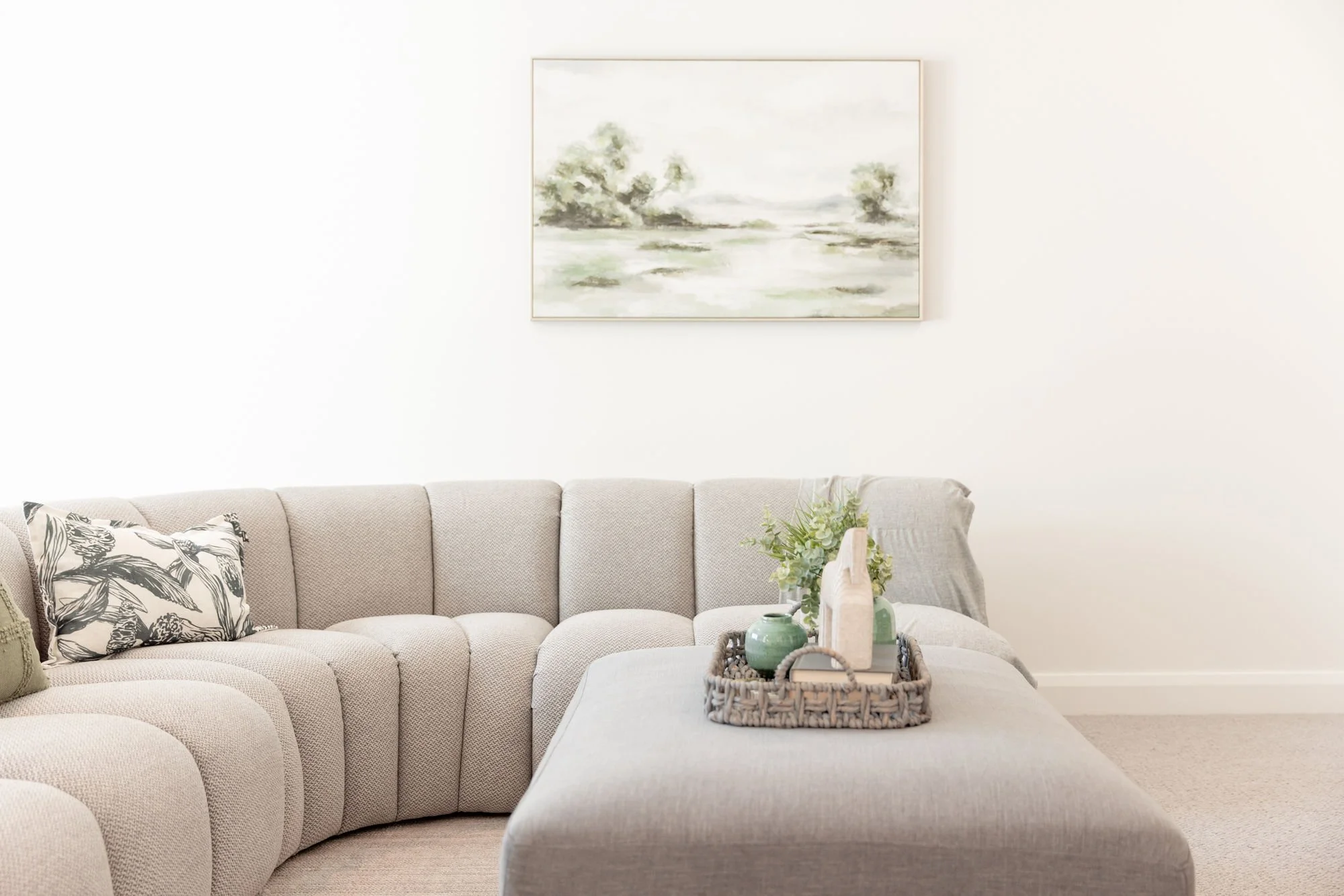 Living room with a light beige sectional sofa, a tray with decorative vases and greenery, artwork of a landscape on the white wall.