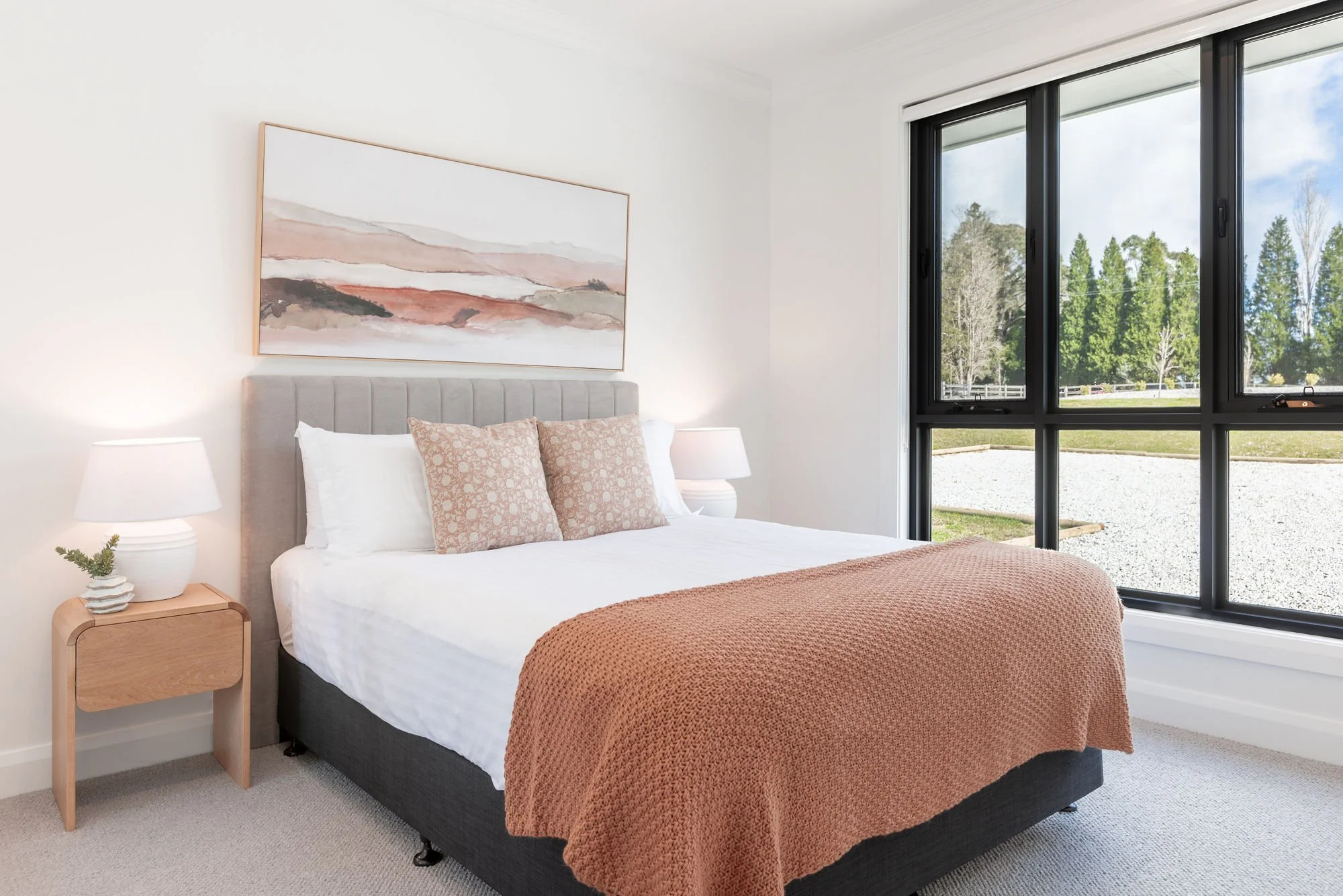 Bedroom with large window, bed with beige pillows, orange blanket, and minimal decor, overlooking green trees outside.