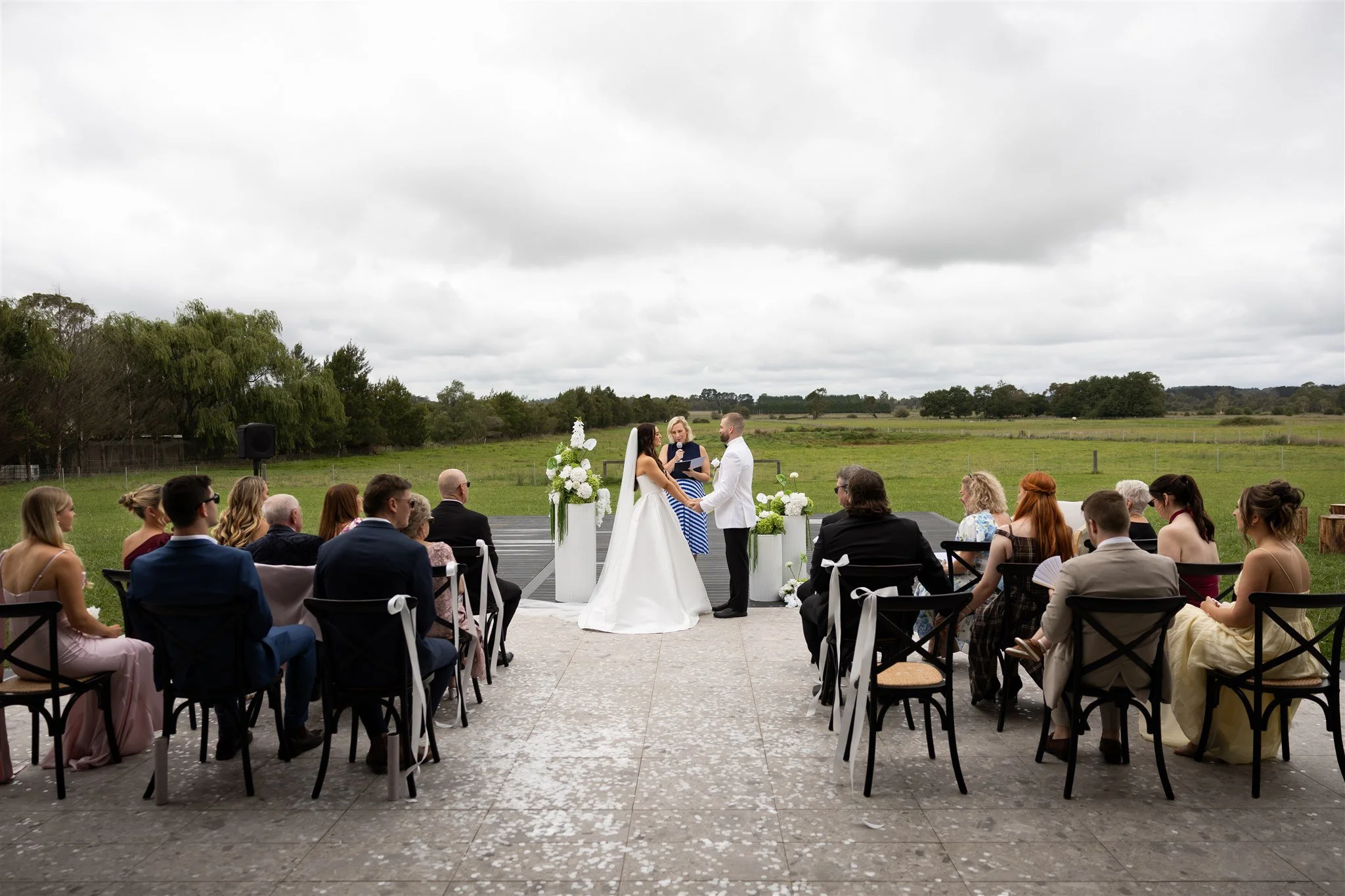 Couple getting married outside with officiant, guests seated in chairs, green fields and trees in the background, overcast sky.