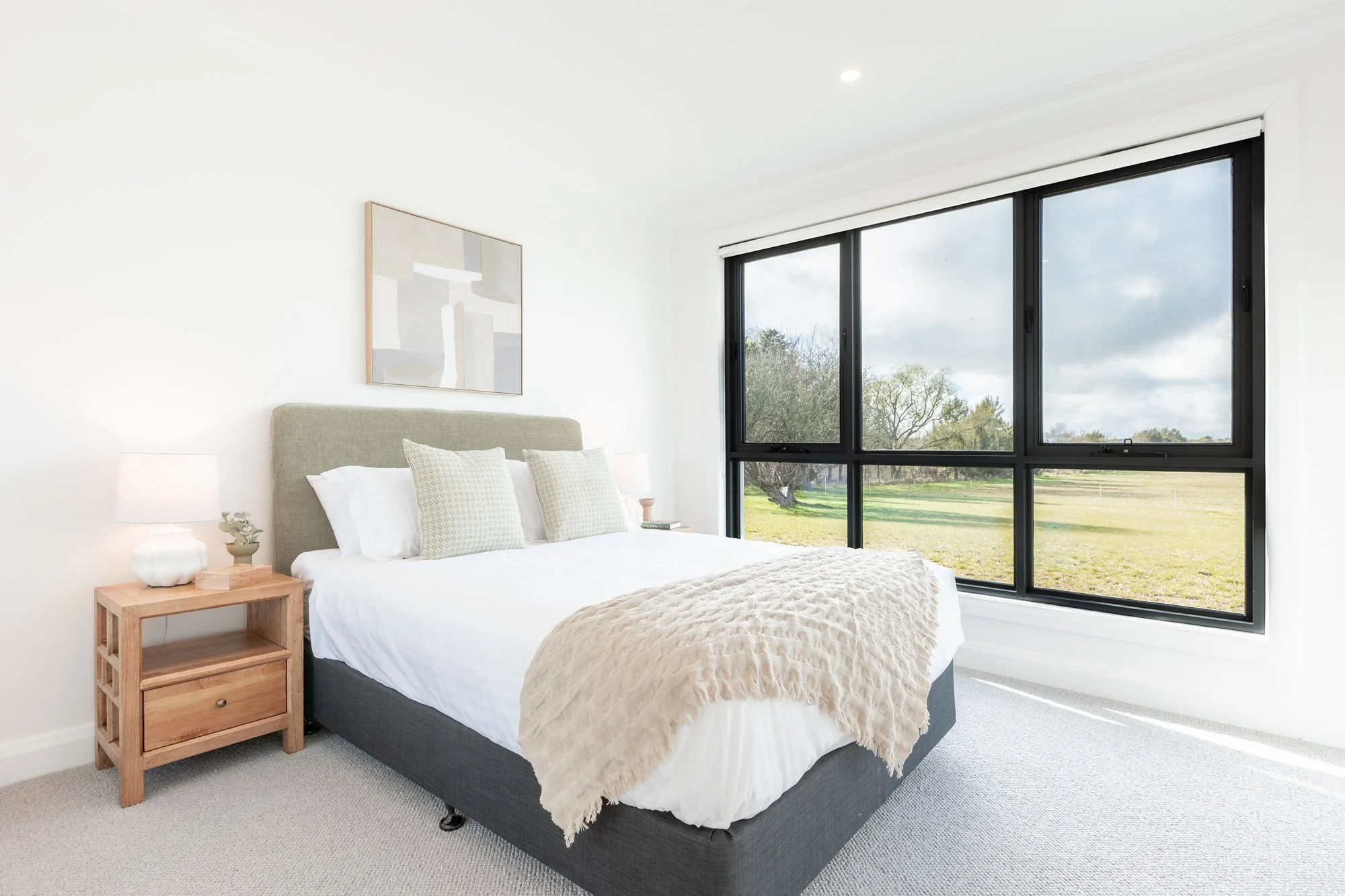 Bright bedroom with large window showing outdoor landscape, bed with white bedding and beige throw, wooden nightstand with lamp and decor, abstract wall art.