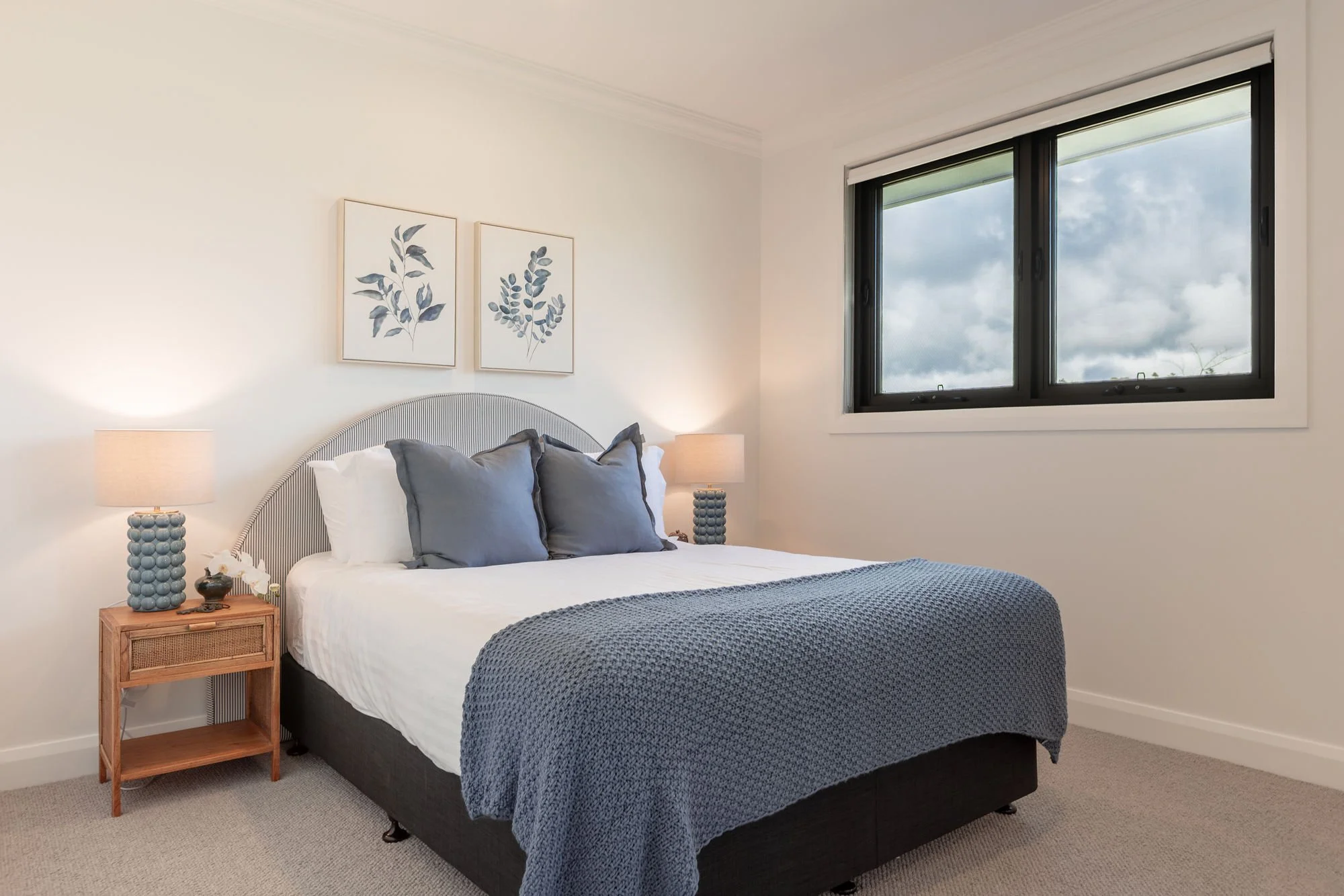 A neatly made bedroom with a bed featuring white sheets, grey pillows, and a textured grey blanket. Two wooden nightstands with lamps and decorative items are on either side of the bed. A window with black framing shows a cloudy sky.