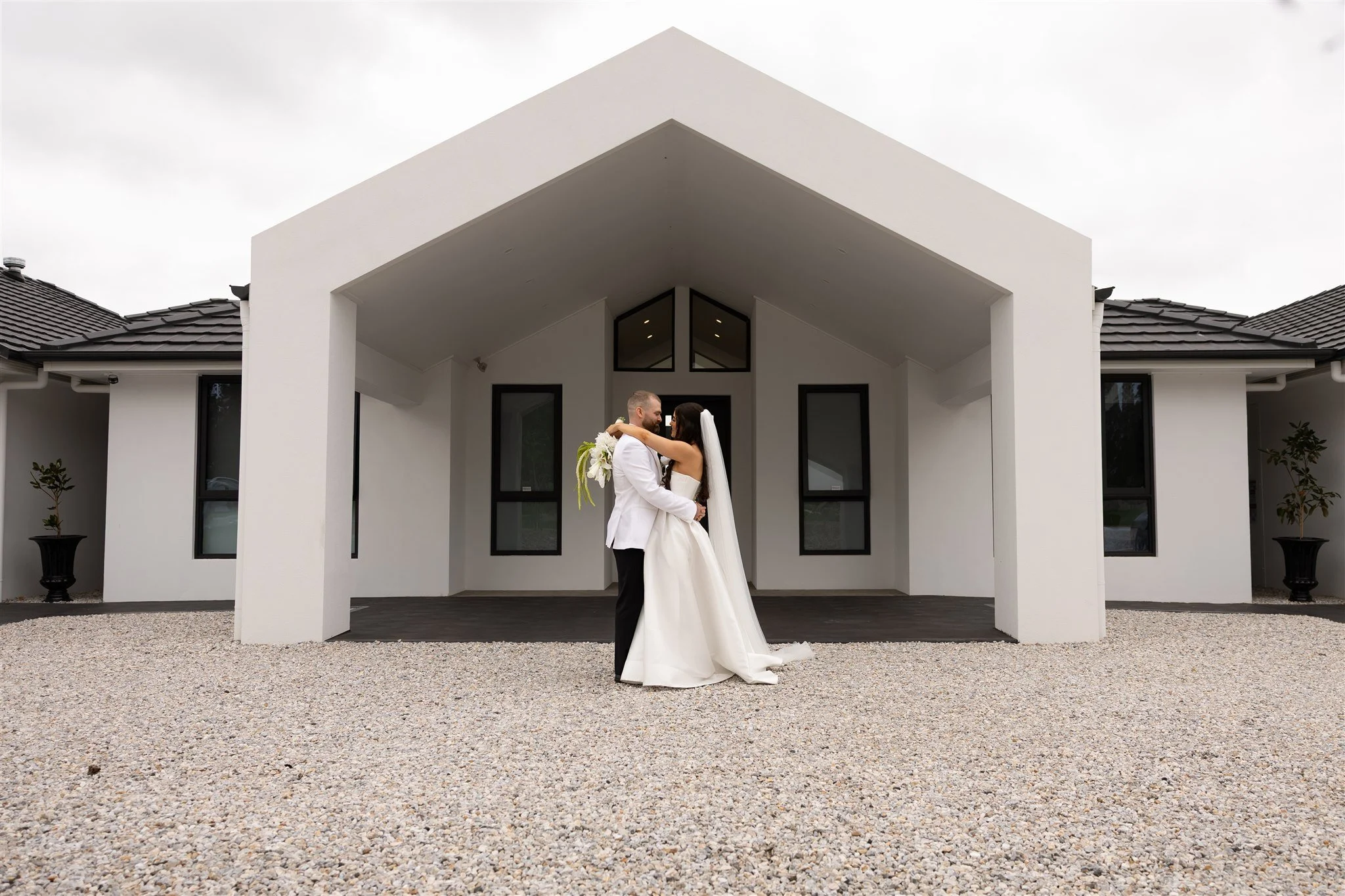 Bride and groom embrace in front of a modern white building on their wedding day.