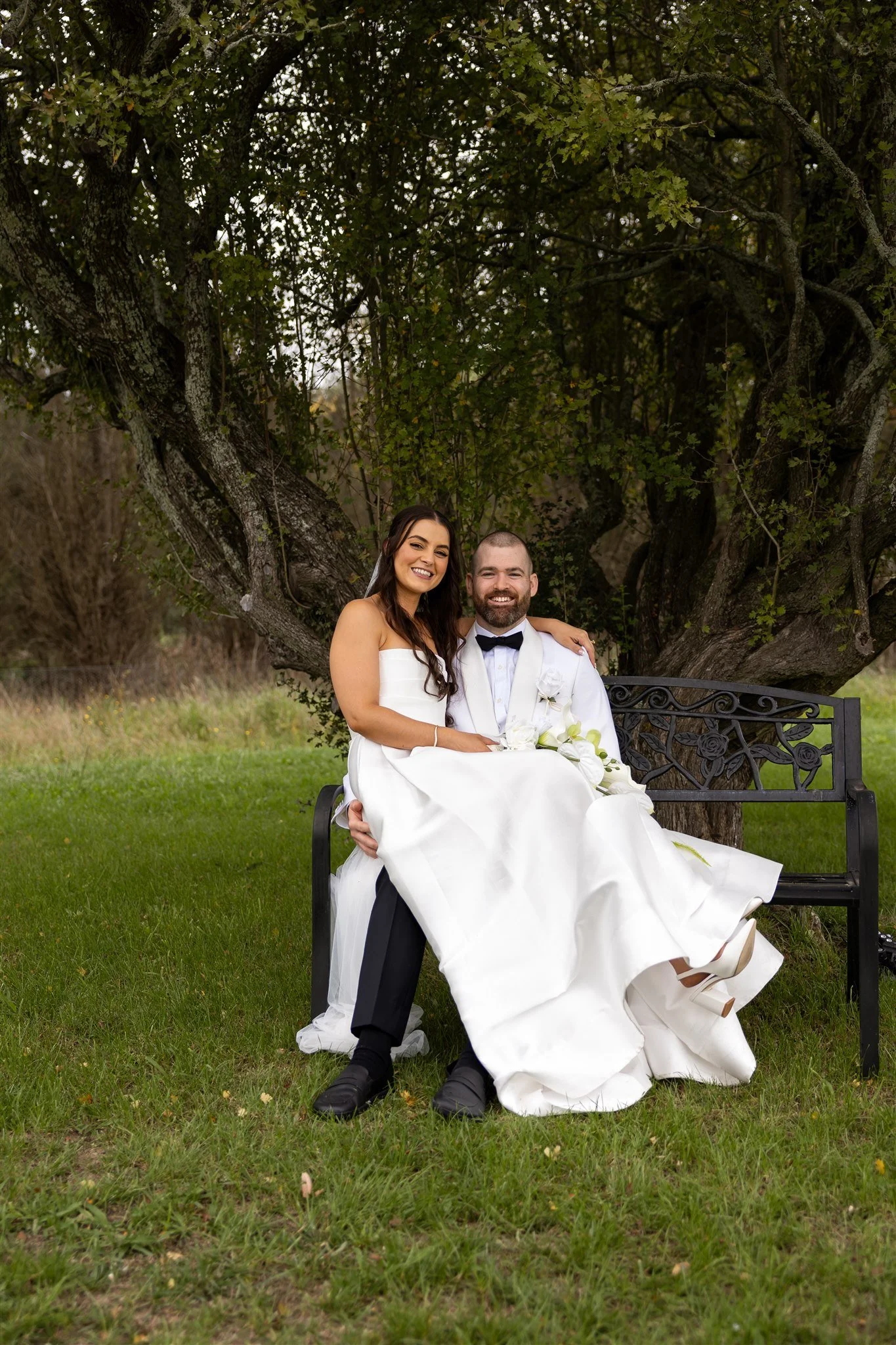 A newlywed couple sitting on a bench under a large tree, smiling for a photo outdoors during daytime. The bride is wearing a white strapless wedding gown and the groom is dressed in a white tuxedo jacket with a black bow tie.