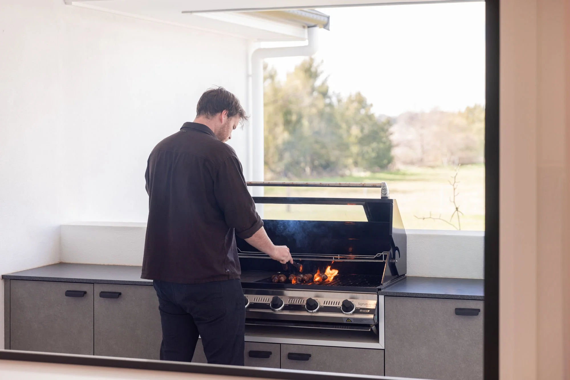 A man grilling food on a barbecue grill outdoors near a large window with trees and grass in the background.