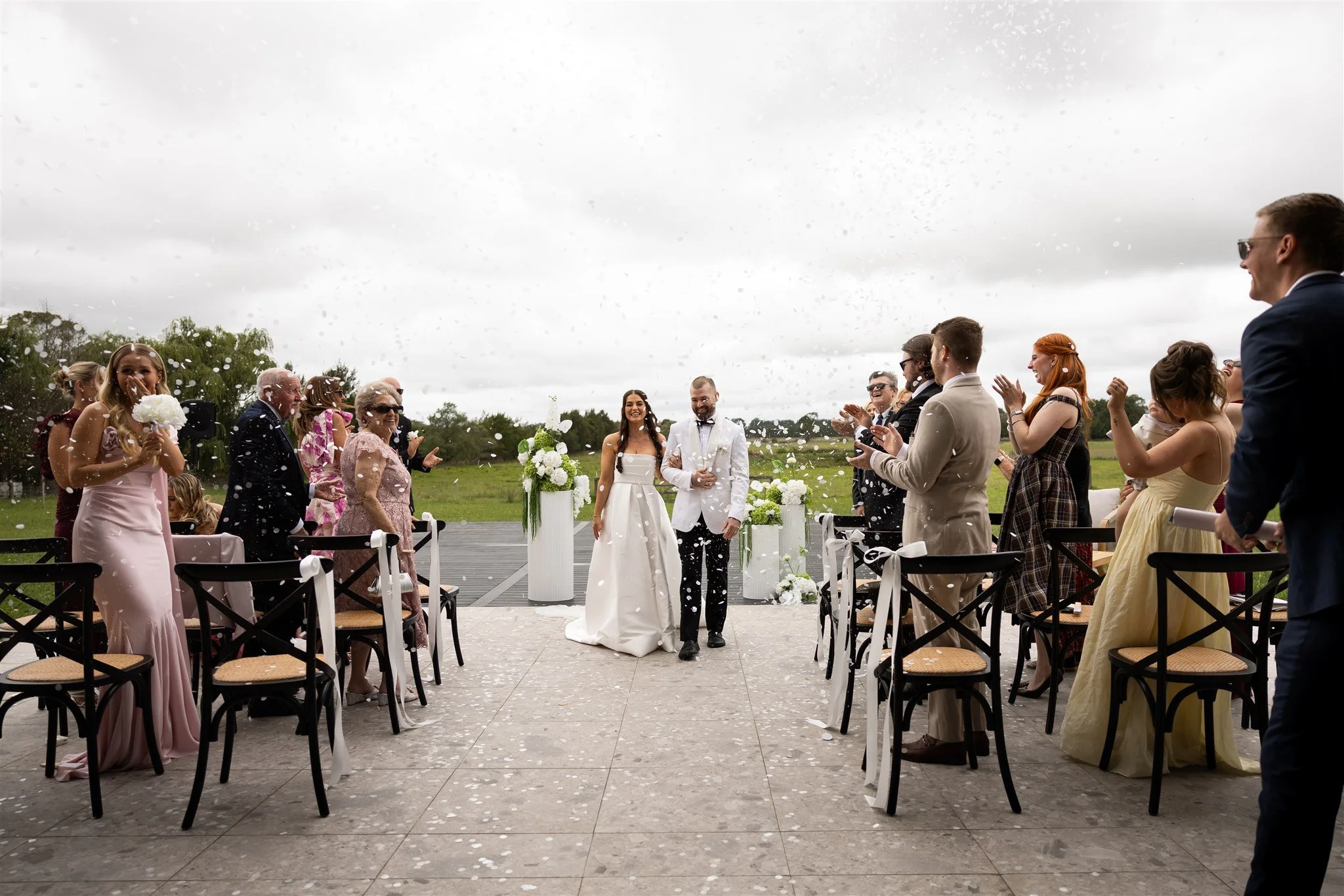 Bride and groom walking down the aisle at outdoor wedding ceremony as guests celebrate with confetti, in a park setting with cloudy sky