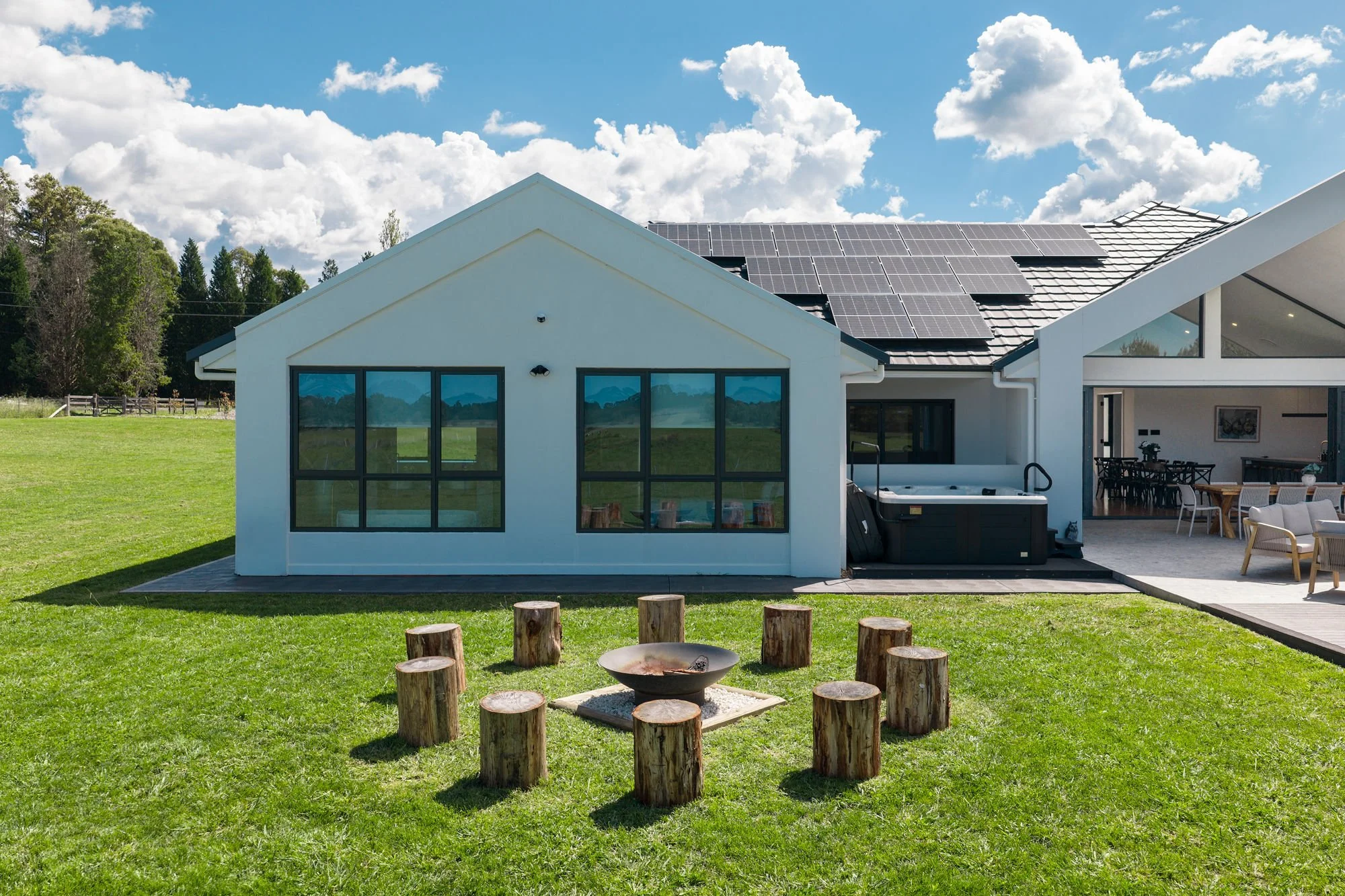 Modern house with large windows, solar panels on the roof, and an outdoor seating area. There is a grassy yard with a fire pit surrounded by wooden logs and a fire bowl in the center. The sky is partly cloudy.