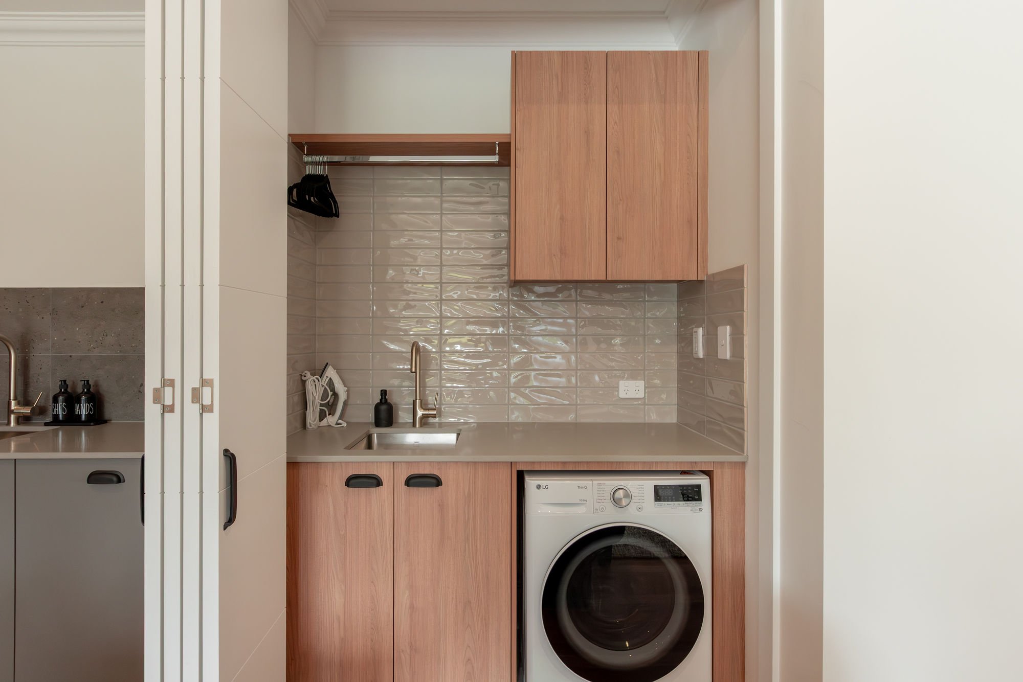 Small kitchen area with a light wood cabinet, a countertop with sink, a black soap dispenser, and a washing machine. There are beige tiled walls and a small white electrical outlet.