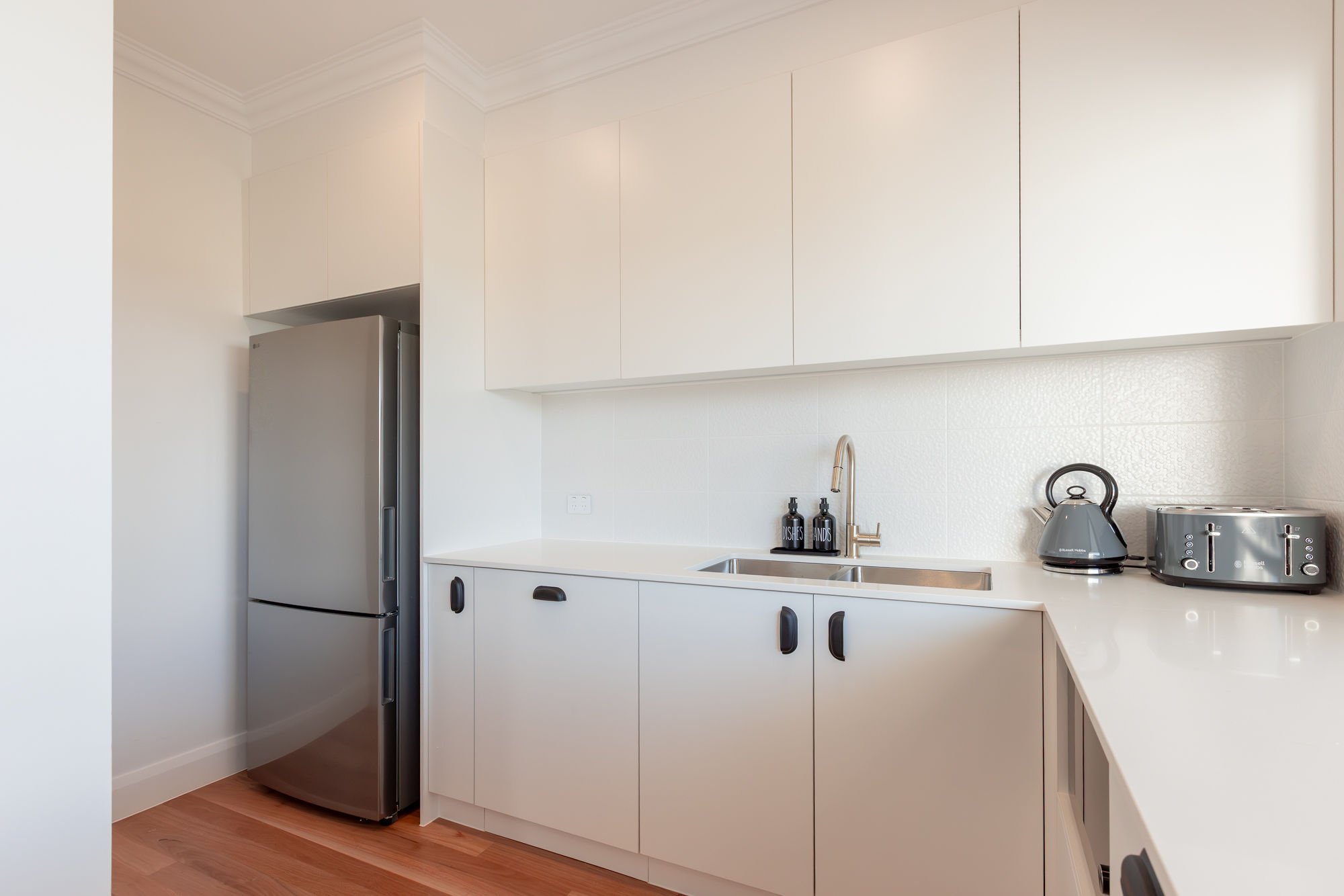 Modern kitchen with white cabinets, stainless steel refrigerator, black and silver toaster and kettle, and black soap dispensers near sink.