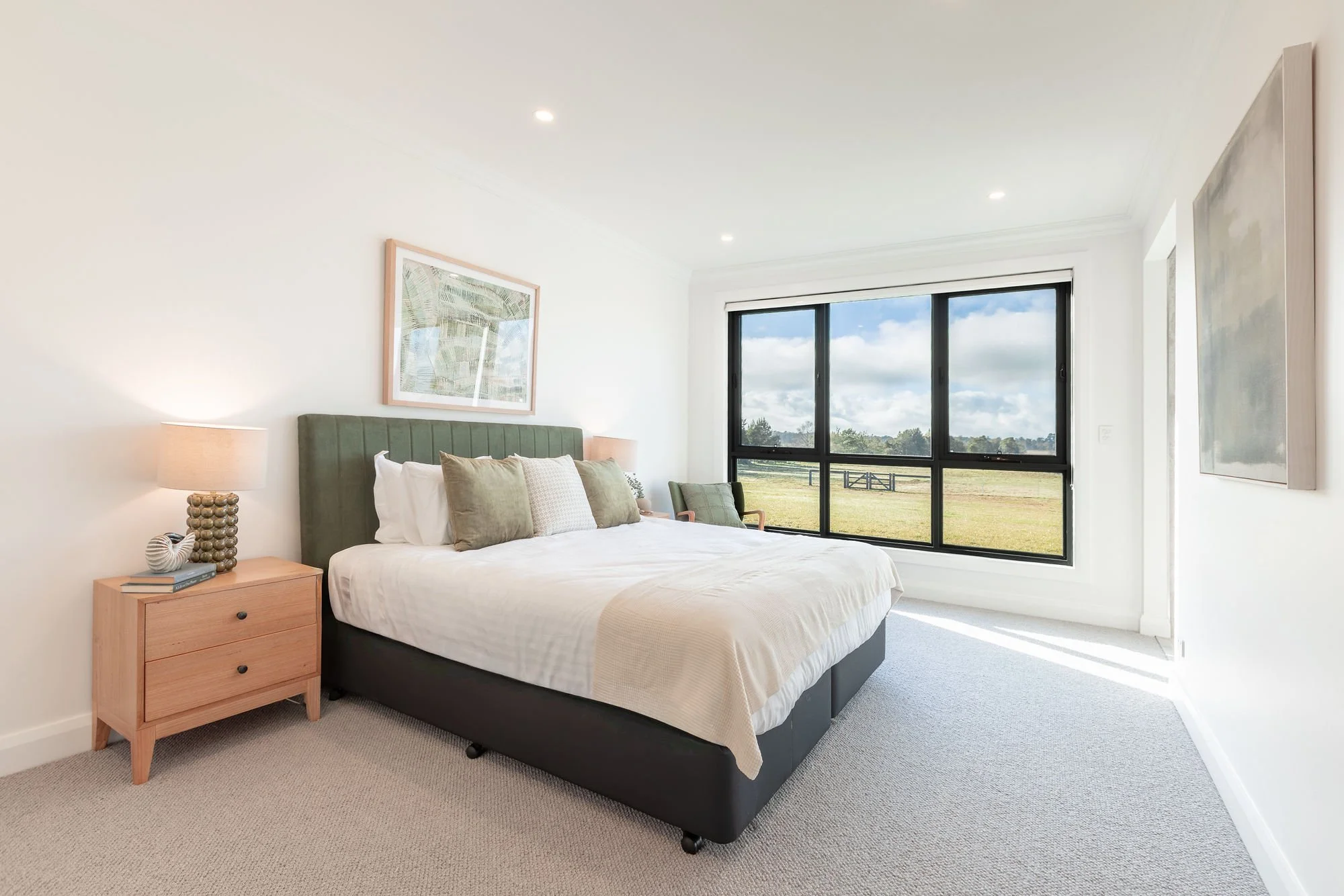 Bright bedroom with a large window showing an outdoor field, a bed with green and white pillows, and a wooden nightstand with a lamp and decorative objects.