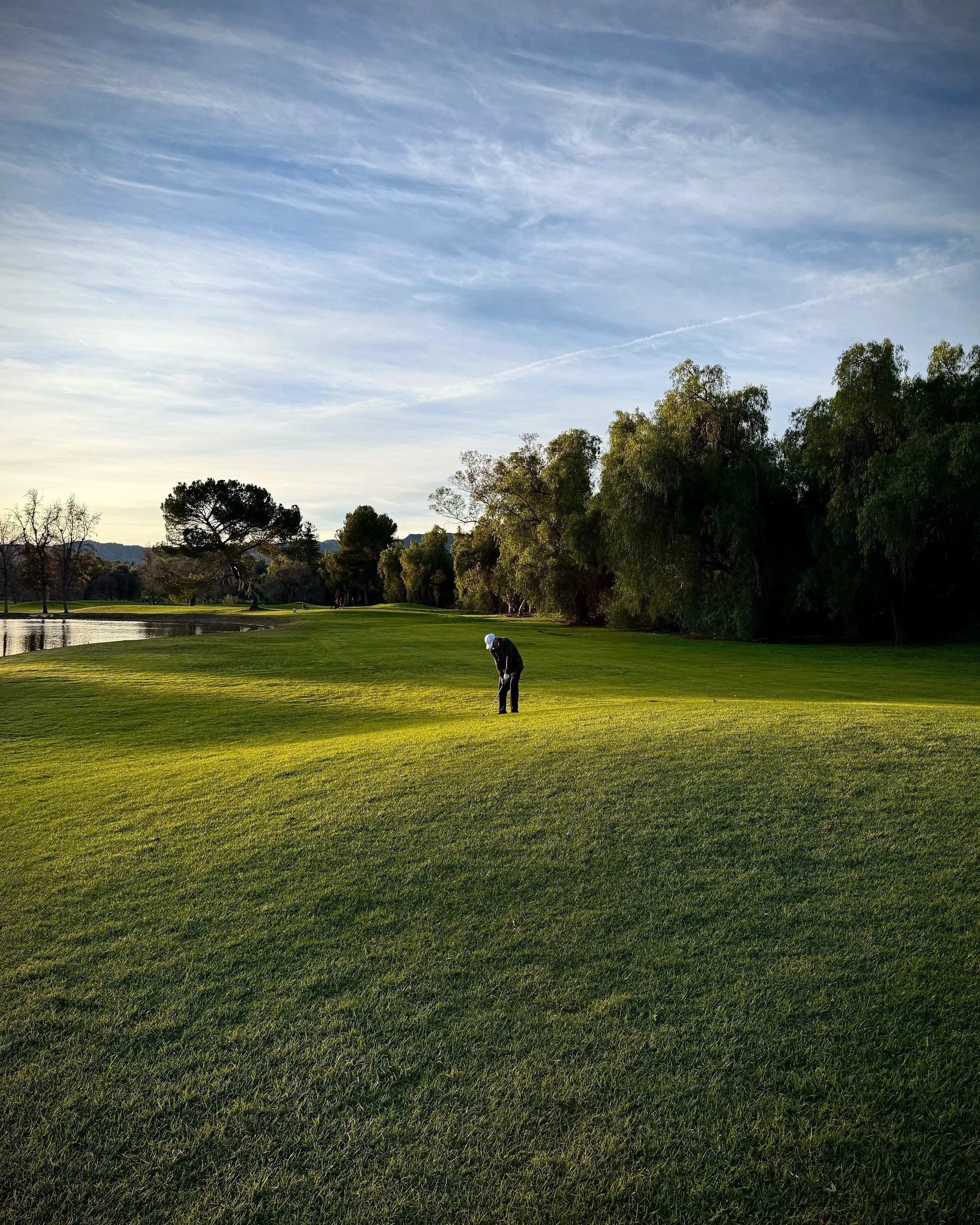 A person in black clothing and white cap playing golf on a lush green course near a water hazard, with trees and a partly cloudy sky in the background.