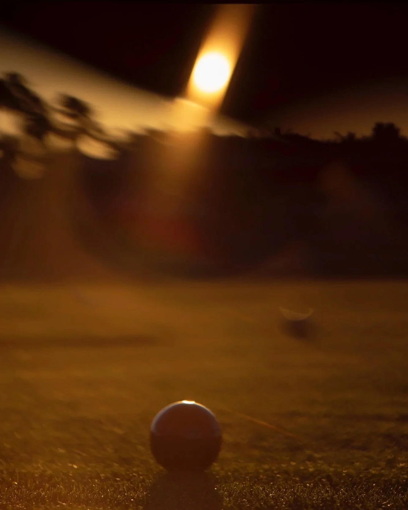 A golf ball on a tee on a golf course at night, with a bright light in the background creating a warm glow.
