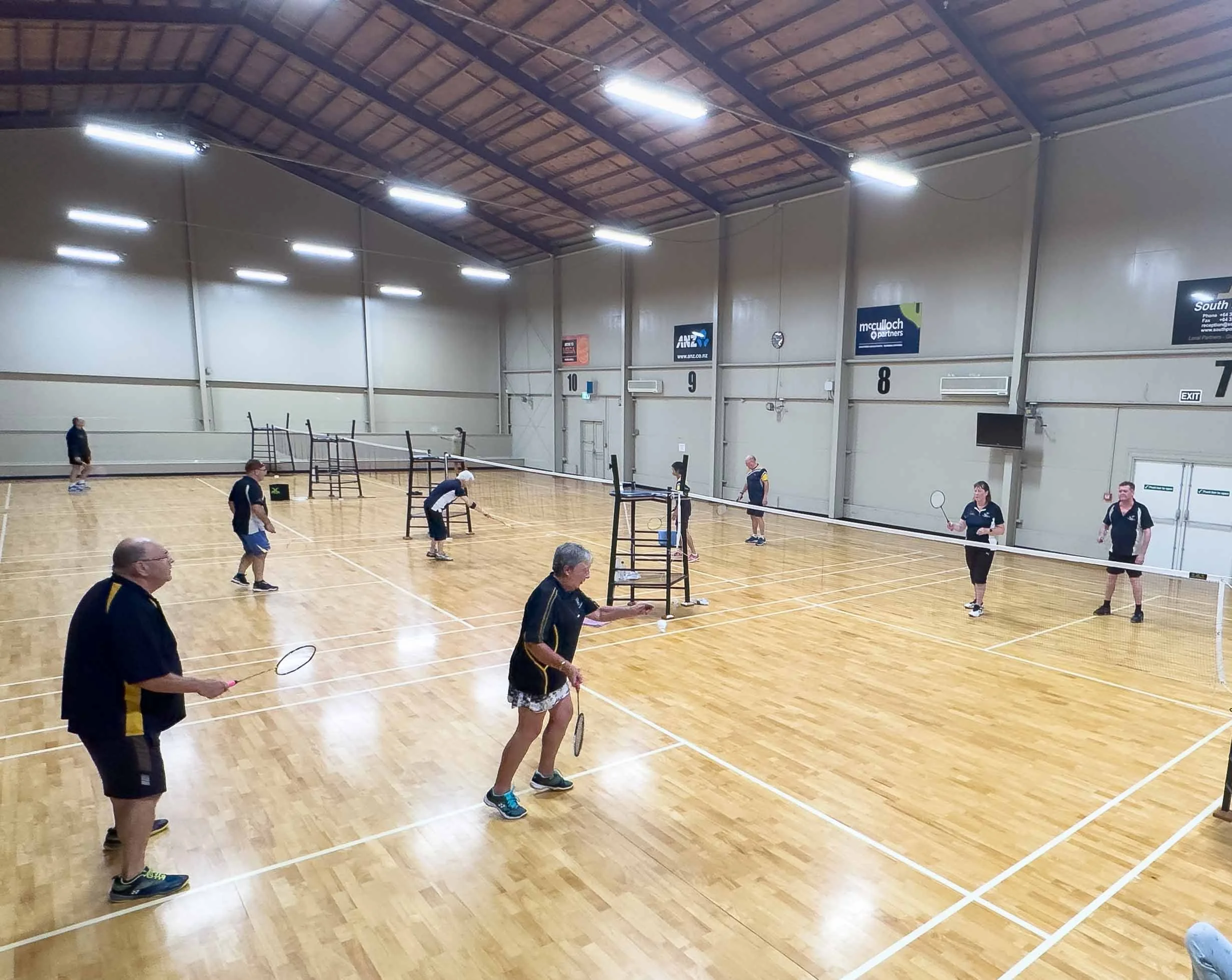 People playing badminton indoors on a large wooden court with 4 nets and 8 players visible.