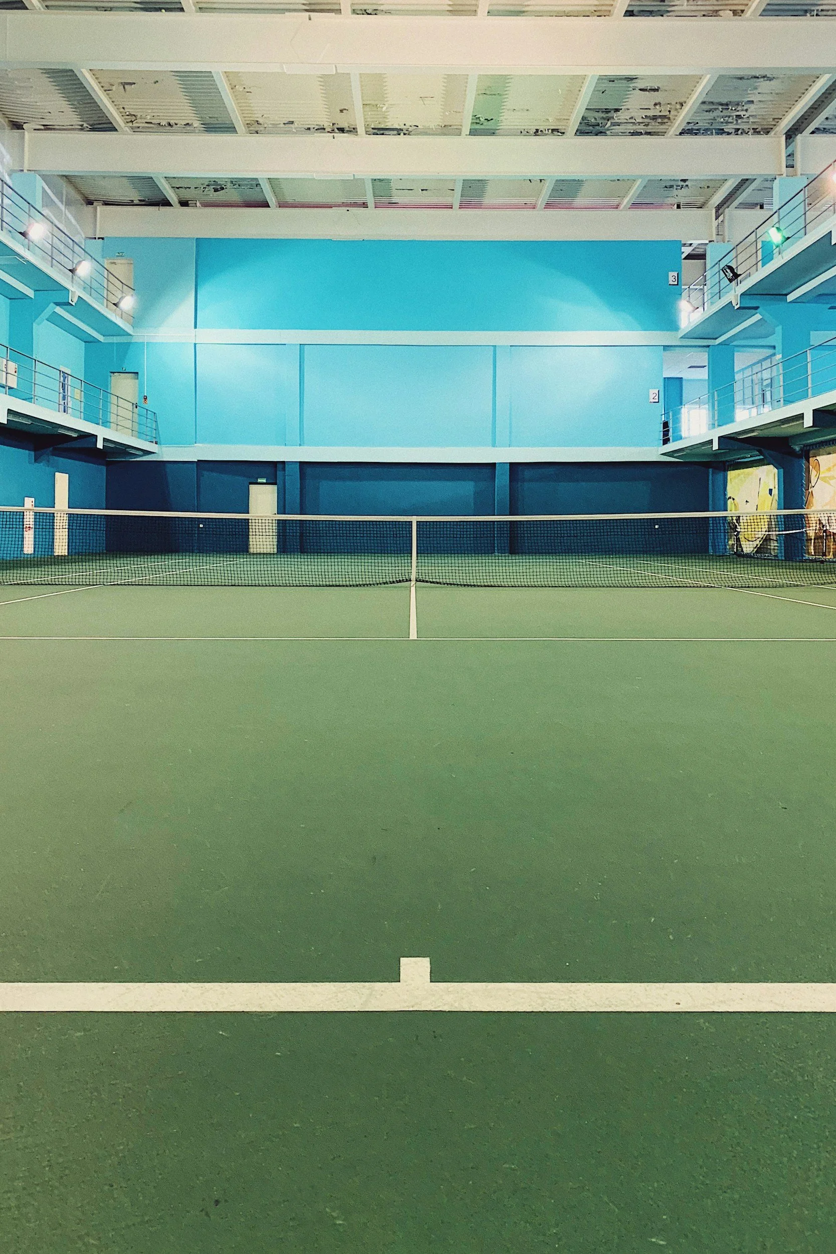 Empty indoor tennis court with green surface, white boundary lines, and blue walls, surrounded by a balcony and railing.