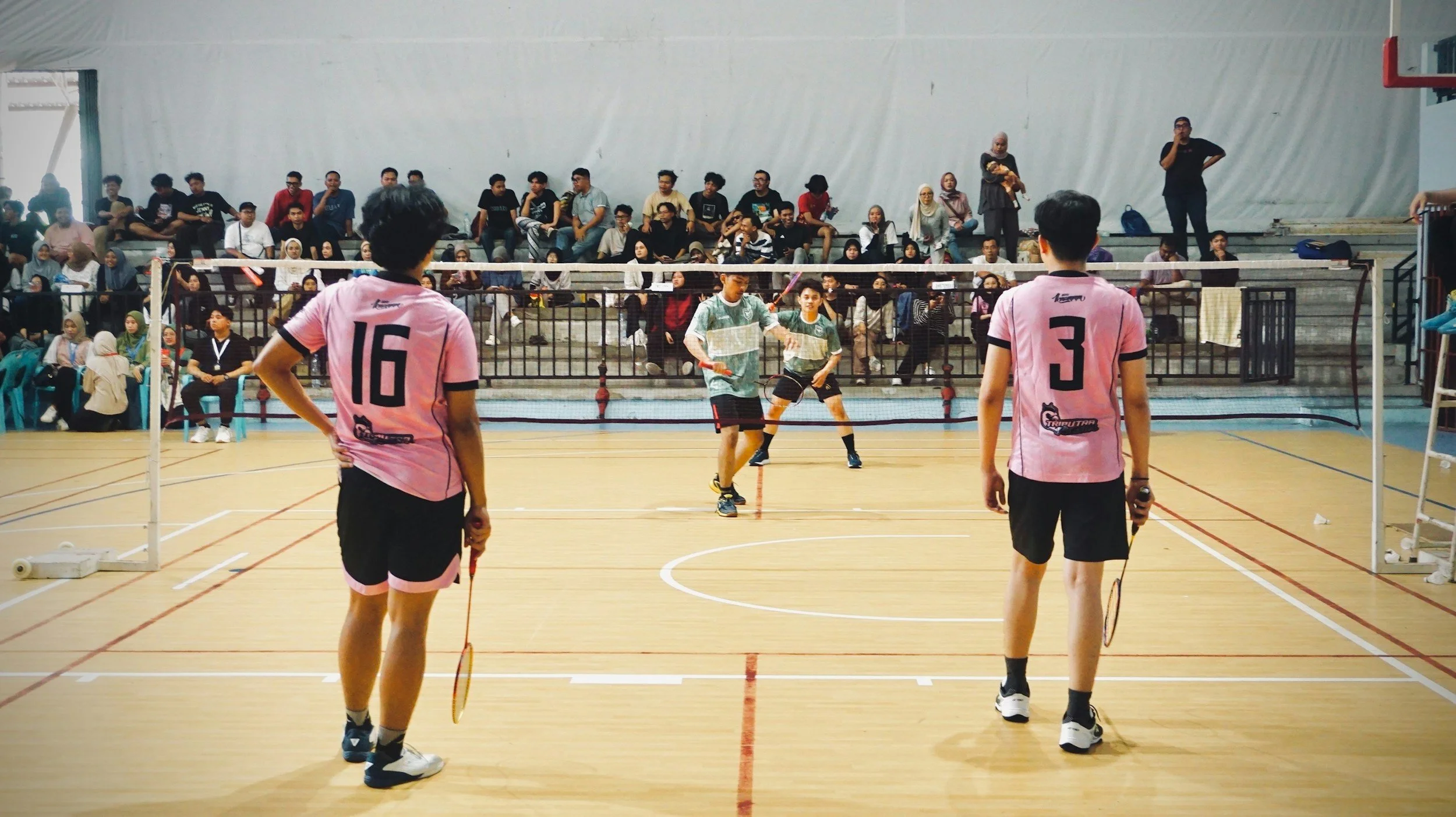 Two young men wearing pink shirts and black shorts playing badminton on an indoor court, with a large audience seated in the bleachers watching.