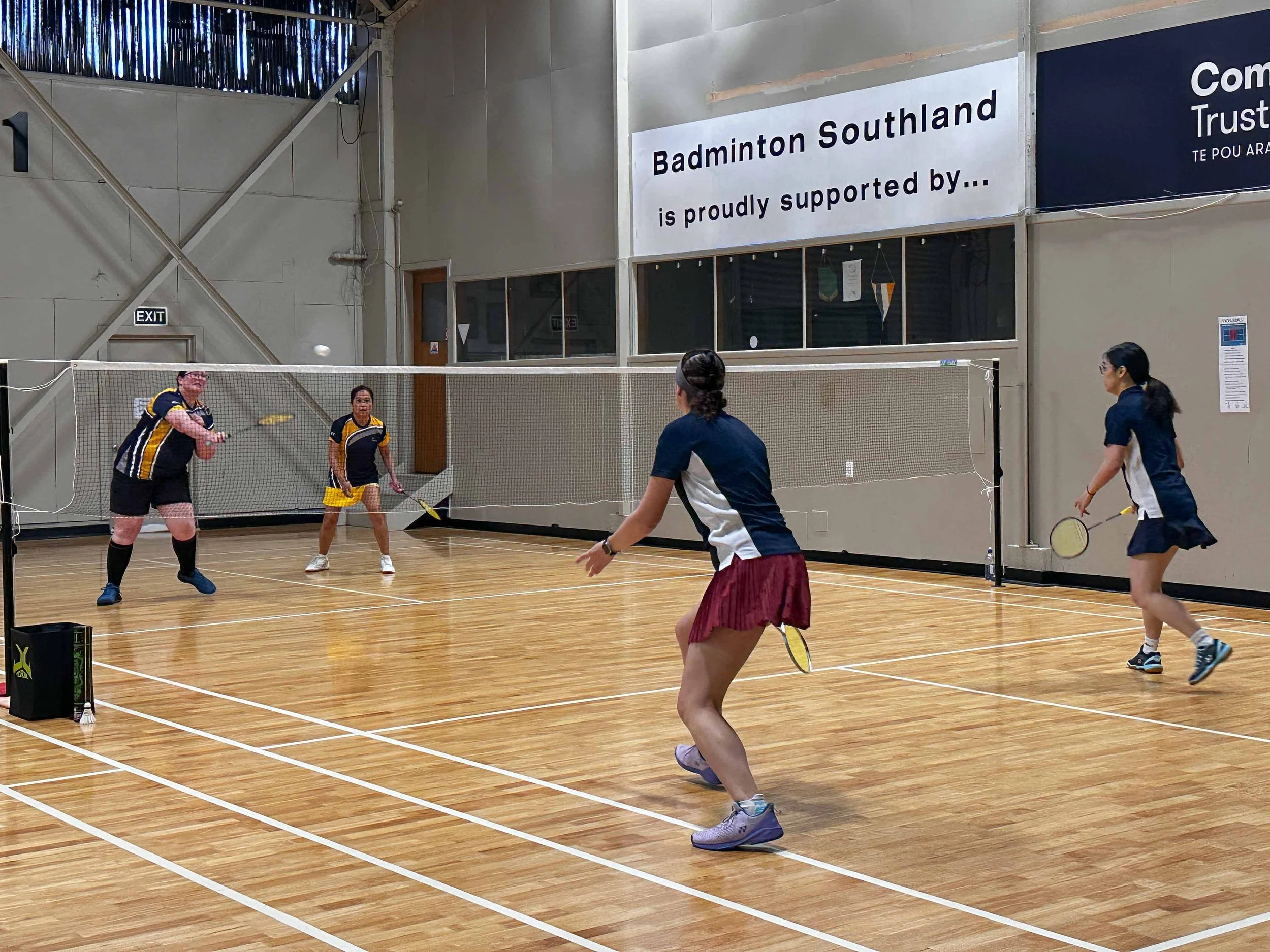 Four women playing badminton inside a sports hall with a wooden floor. Two women are on each side of the net, with one woman in the foreground preparing to hit the shuttlecock and the others ready for the game. There is a banner reading 'Badminton So