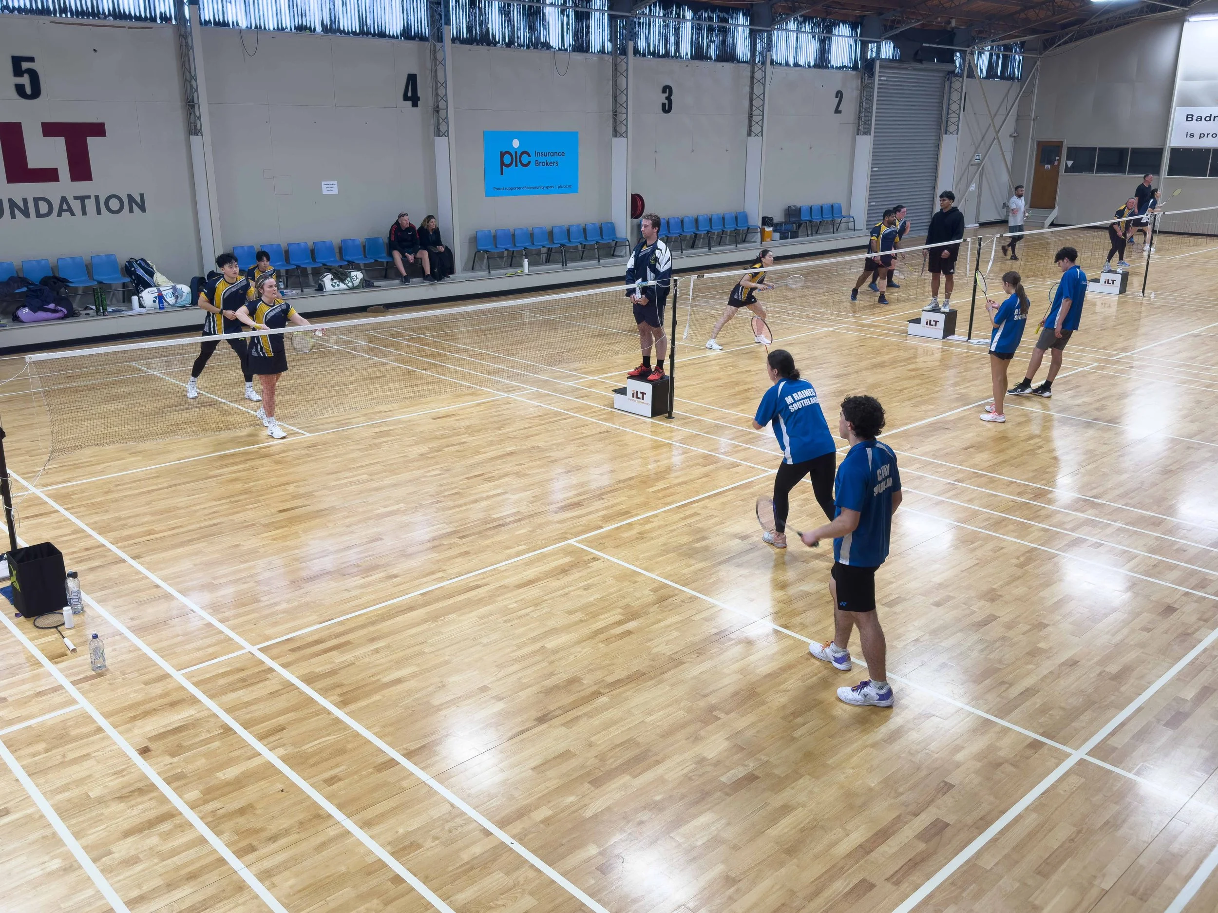 Indoor badminton court with players practicing and coaches observing, notable logos on wall and court side.