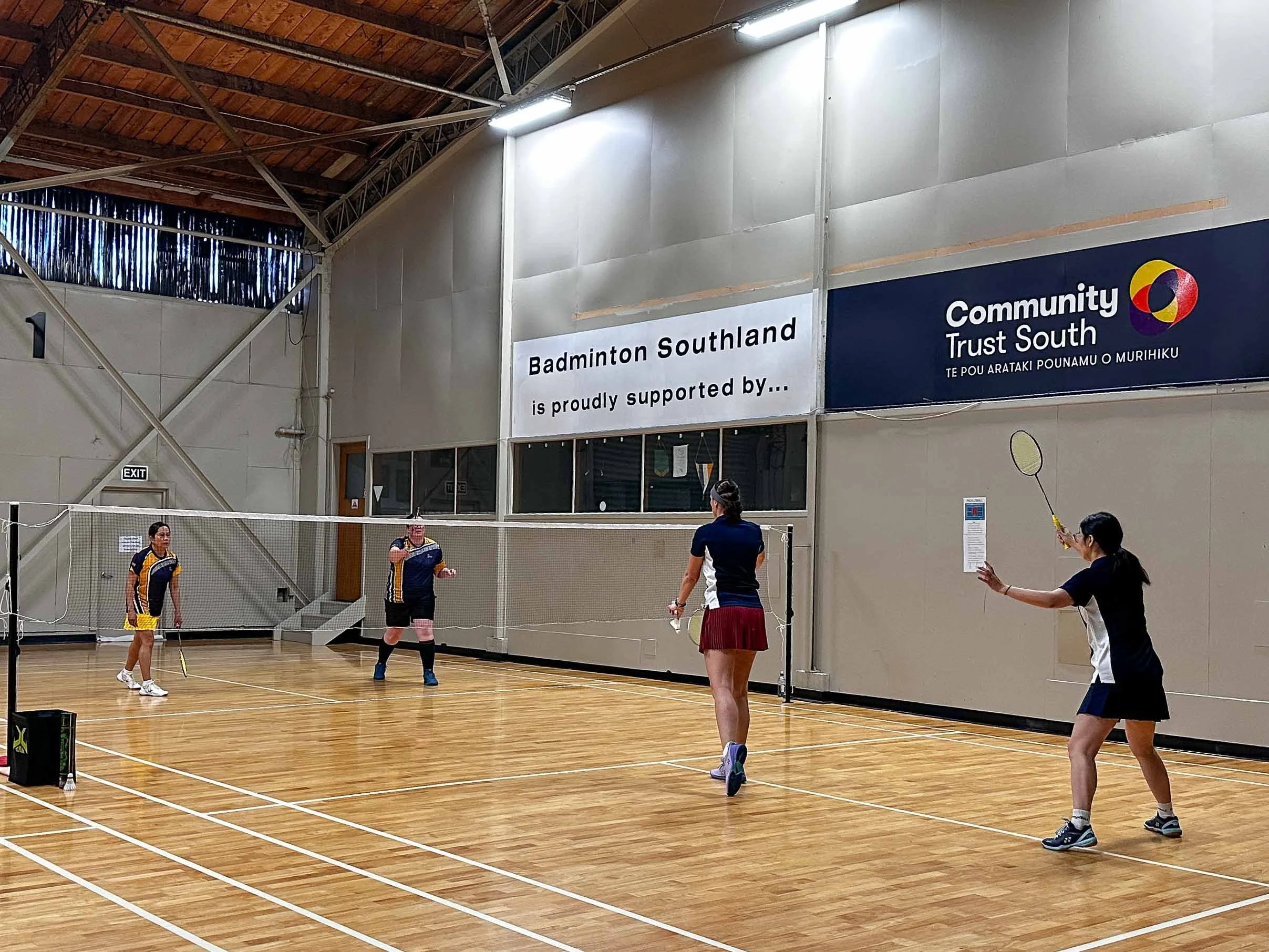 Four women playing badminton on an indoor court with a wooden floor, a net, and banners on the wall supporting Badminton Southland and Community Trust South.